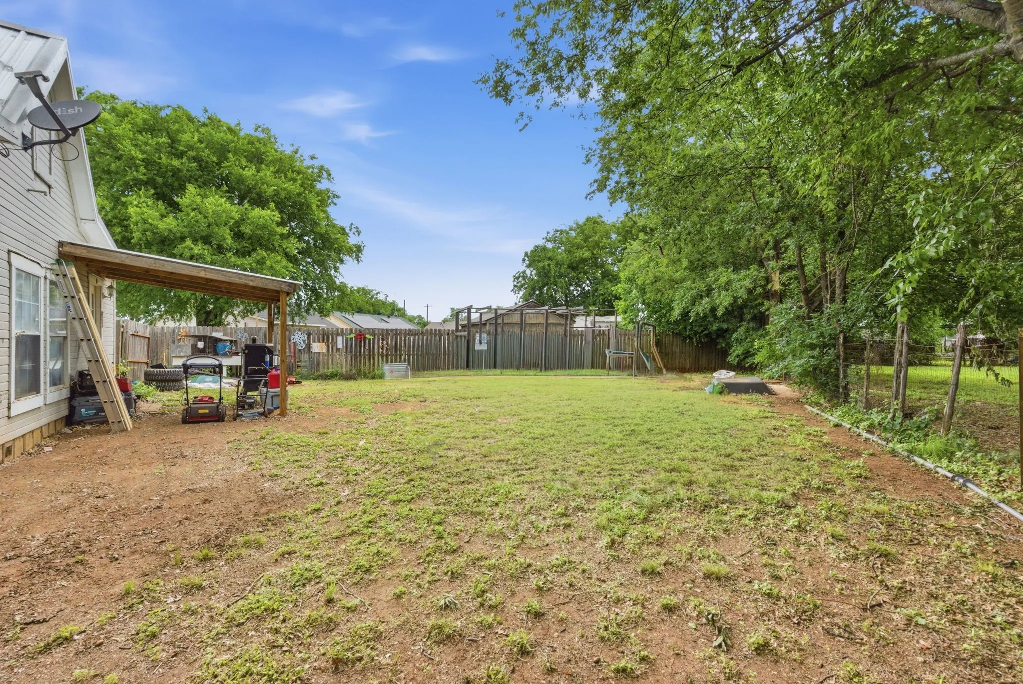 Expansive backyard featuring a partial wooden privacy fence, mature perimeter trees, and a covered patio area with a slatted wood roof