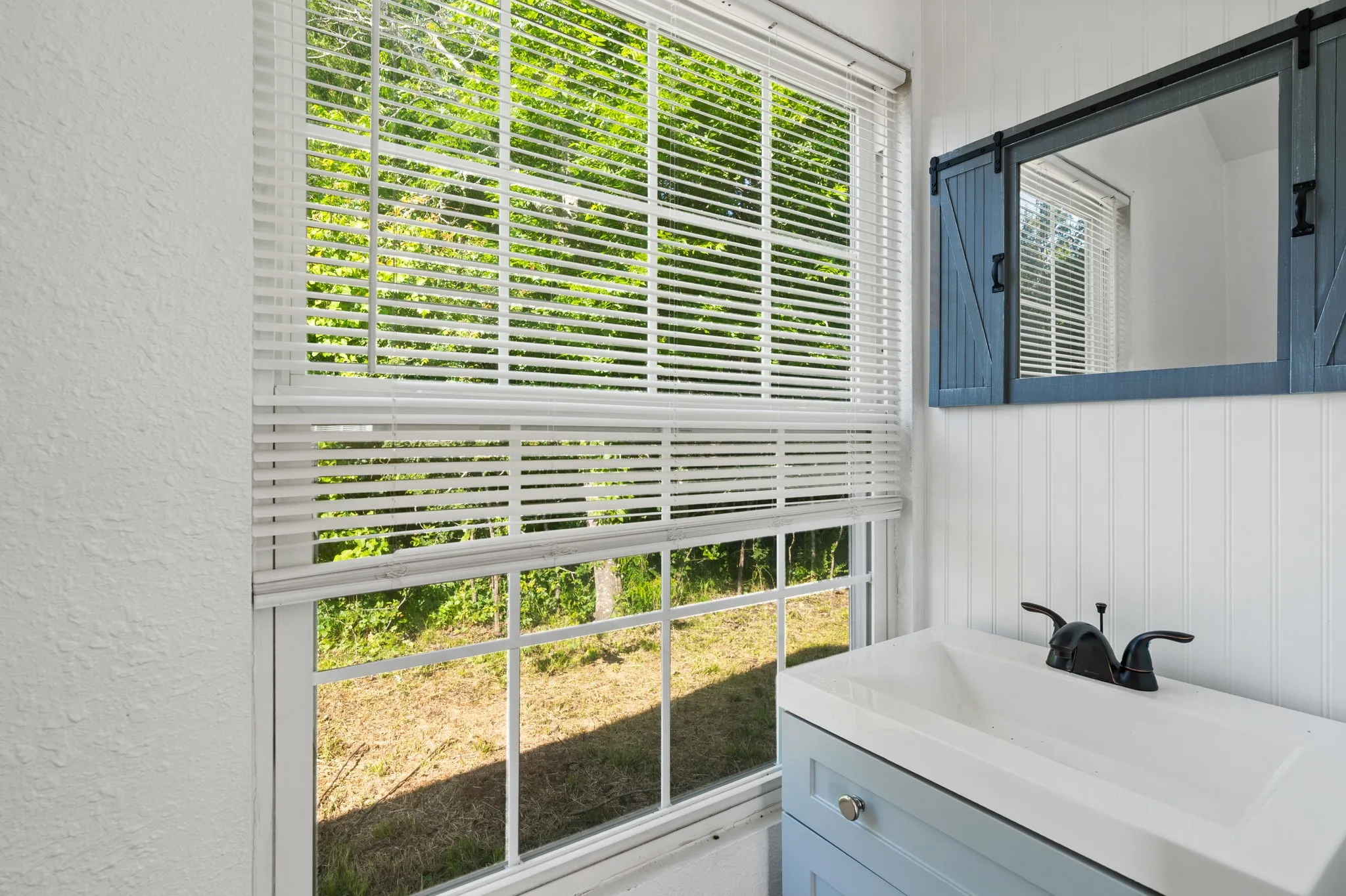 Secondary Bathroom featuring a window with horizontal blinds, a light blue vanity, an integrated sink, a dark bronze faucet, and a barn door style mirror
