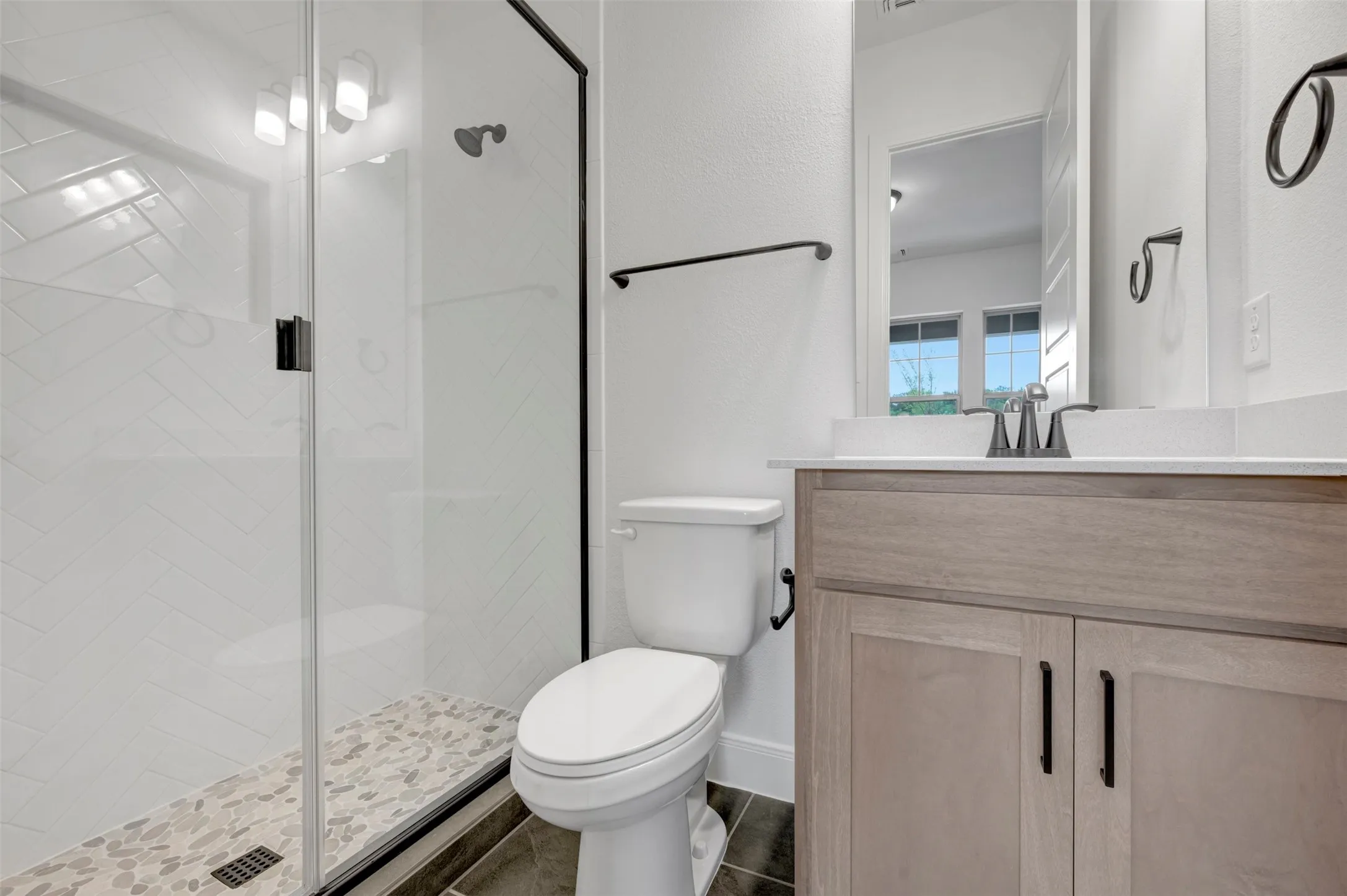 Glass-enclosed shower with subway tile surround and pebble-stone flooring, wood-finish vanity with countertop, and dark-toned floor tile