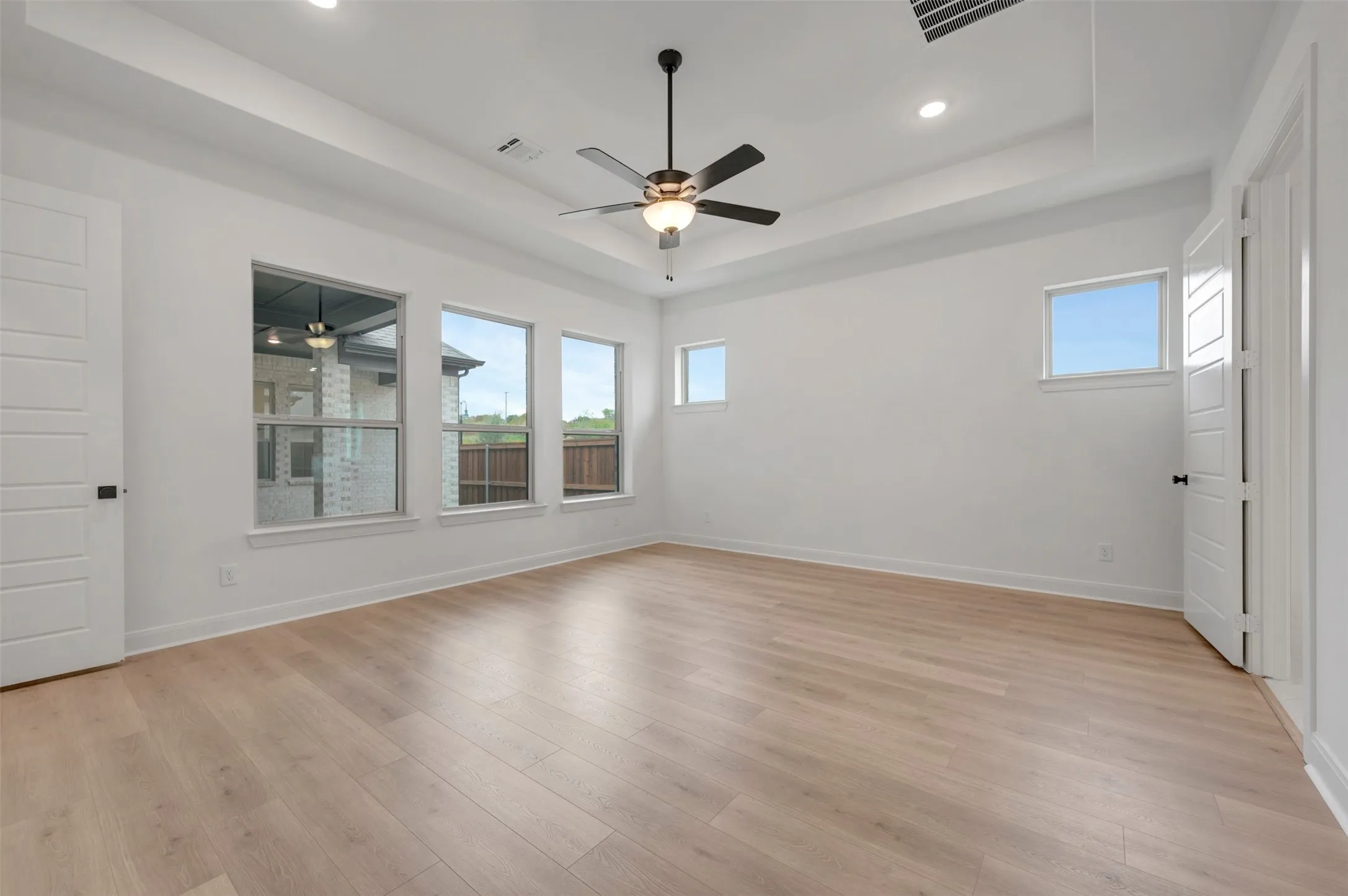Spacious room featuring wood-finish flooring, a tray ceiling, multiple windows, and a ceiling fan with integrated lighting