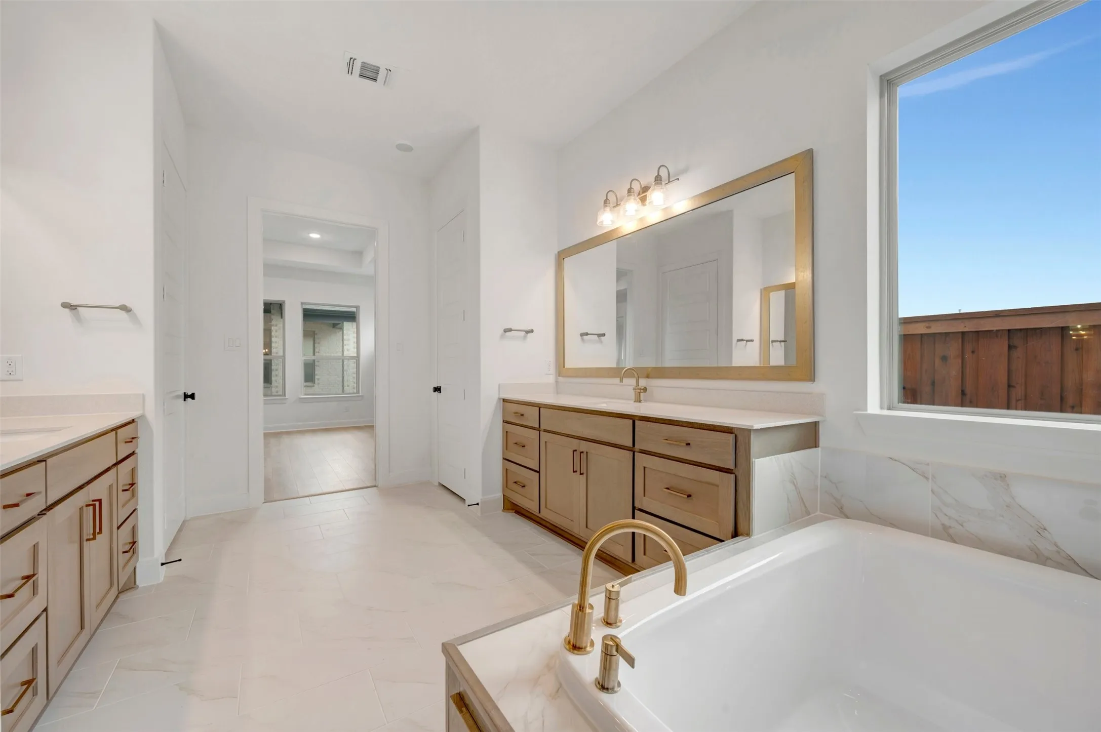 Dual vanities with light wood-finish cabinetry, a large soaking tub with brass fixtures, a wide window, and a spacious mirror with overhead lighting