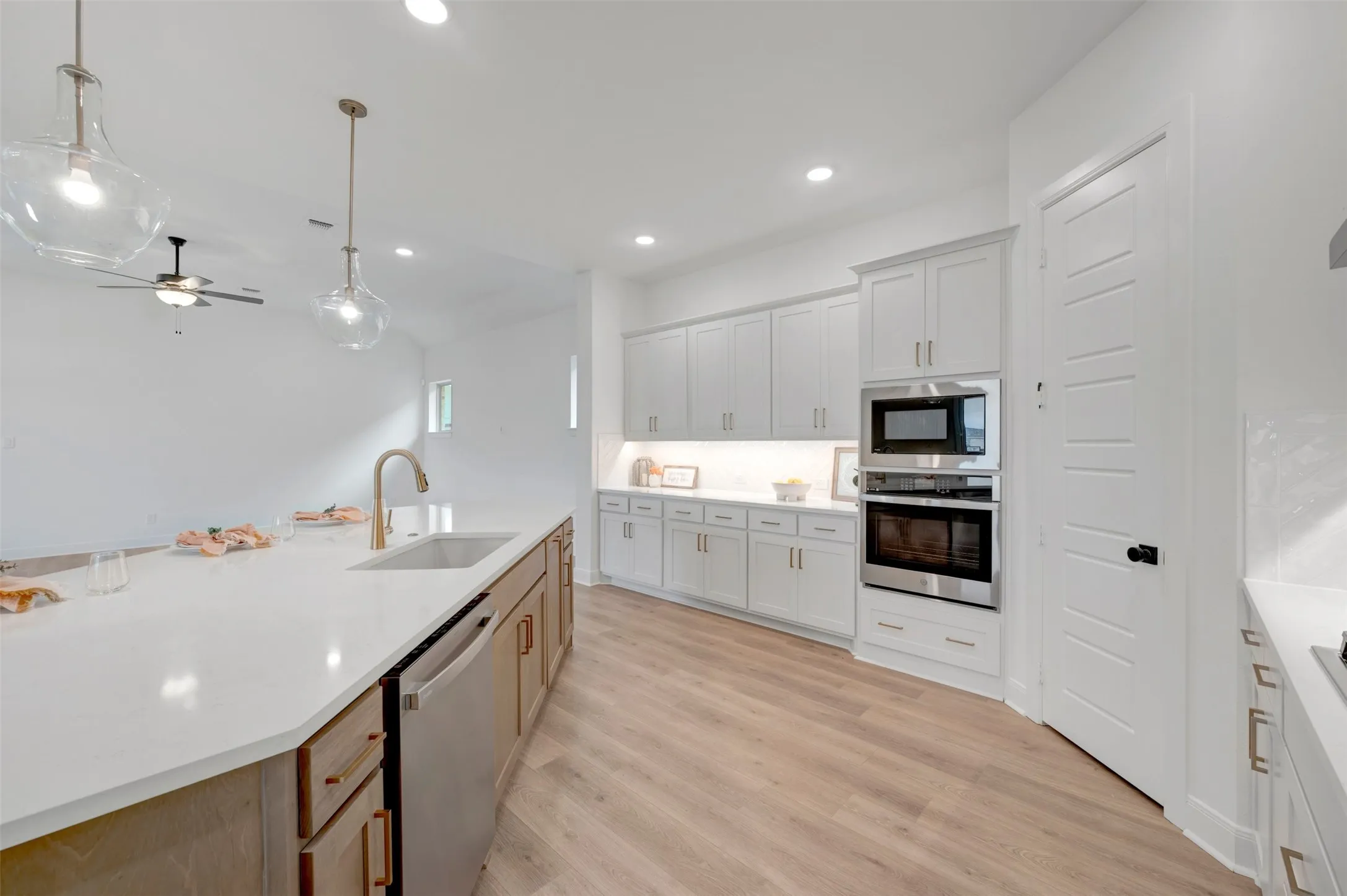 Spacious kitchen featuring an expansive island with an undermount sink, built-in stainless steel appliances, light-toned wood-finish flooring, and white cabinetry with under-cabinet lighting