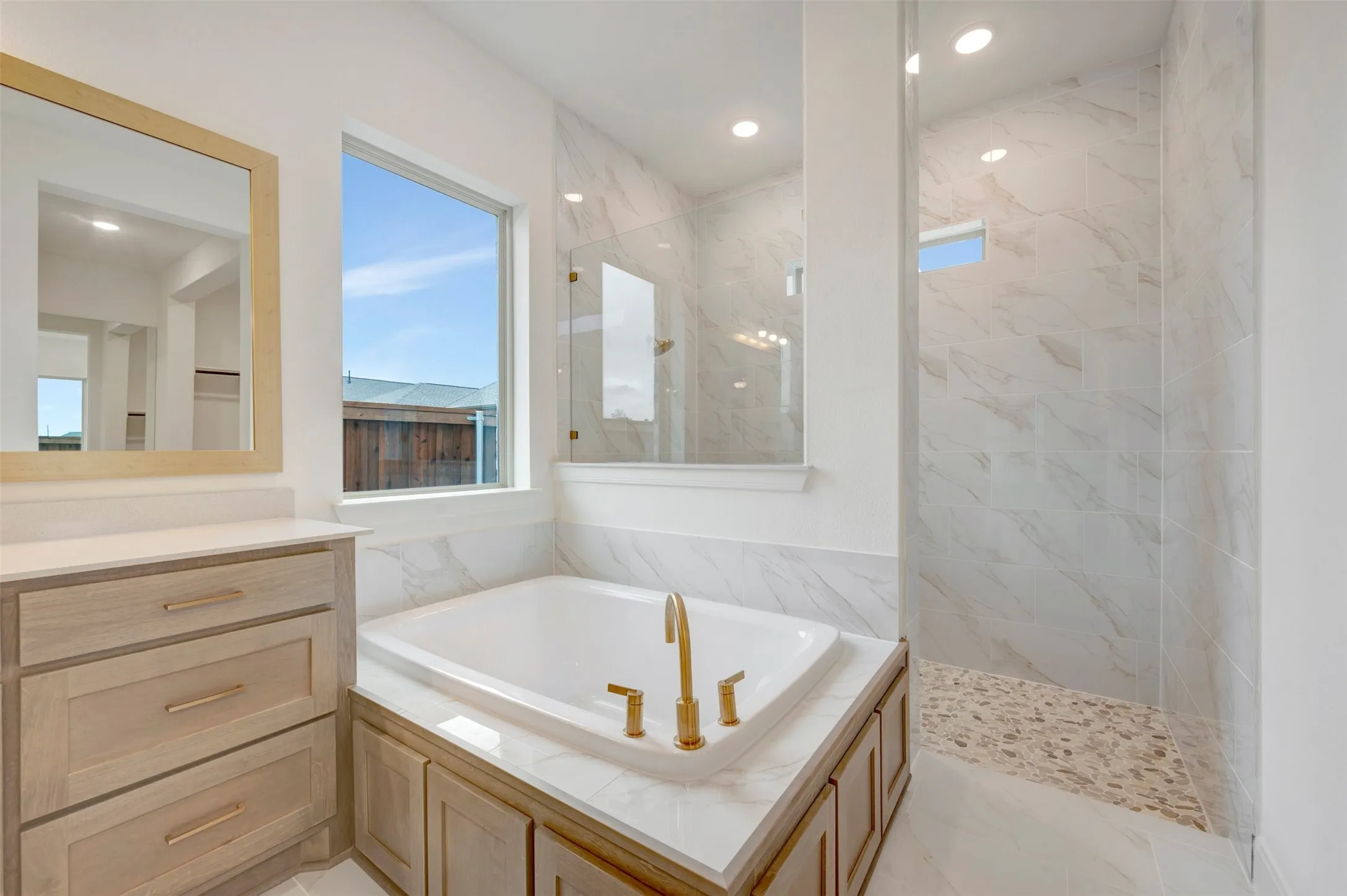 Built-in soaking tub with gold-tone fixtures, walk-in shower with natural stone tile, large window, and custom wood-finish cabinetry