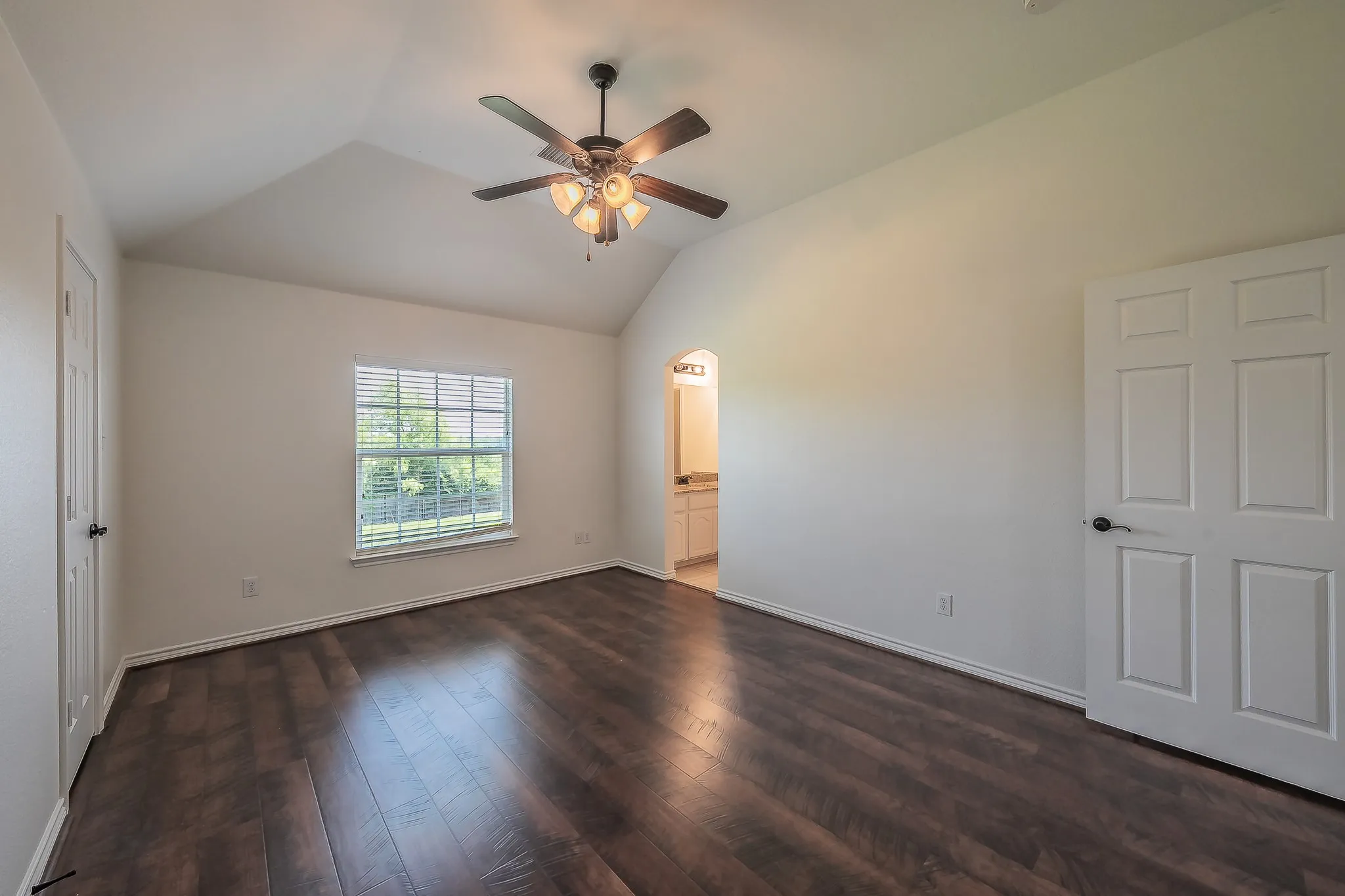 Bright secondary bedroom with vaulted ceiling, ceiling fan, hardwood floors, picture window with green views, and an arched doorway leading to the private ensuite bath.