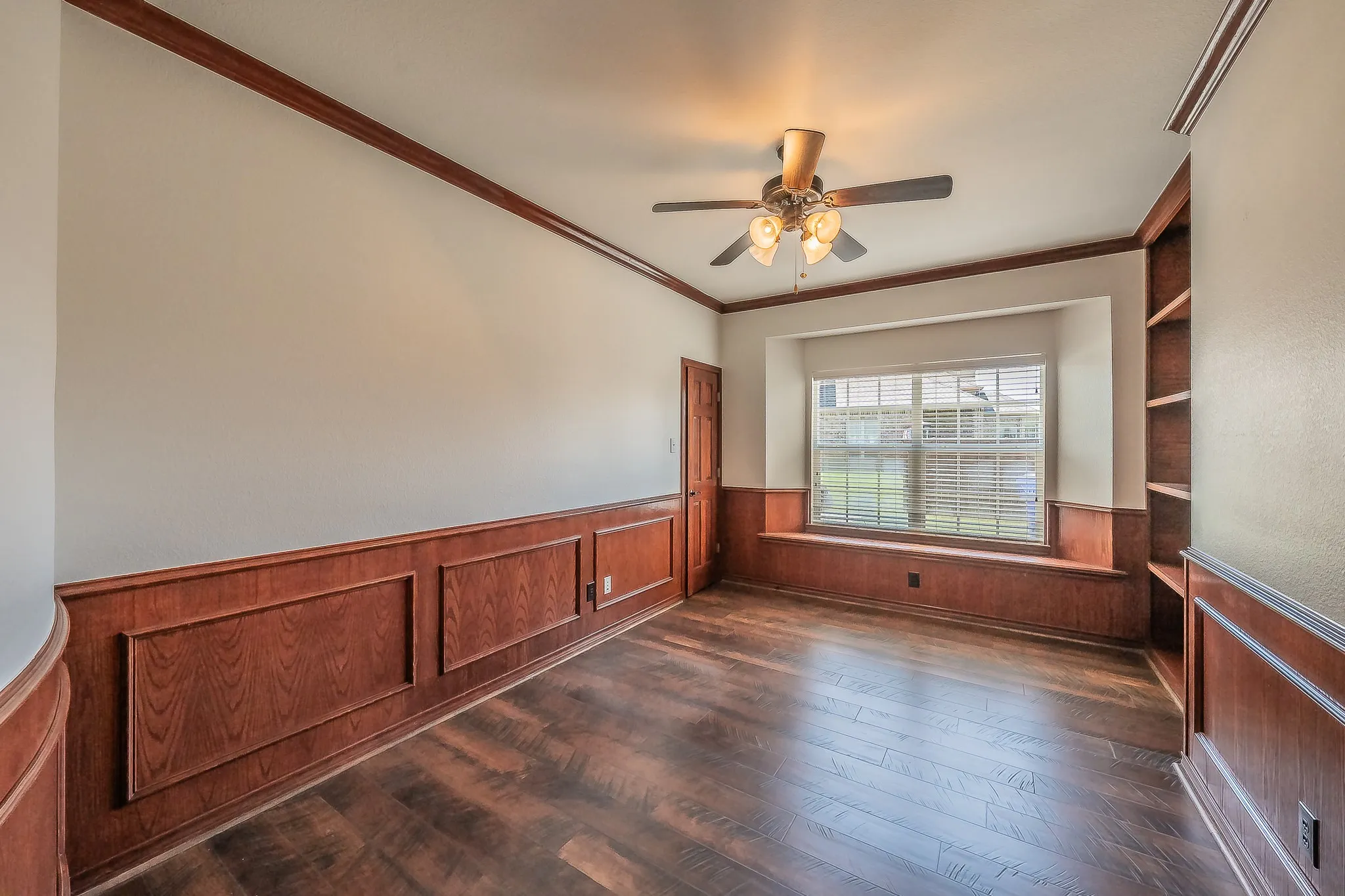 Elegant first-floor study featuring rich wood wainscoting, built-in bookshelves flanking a bay window seat, ceiling fan with light, and warm hardwood floors — an ideal home office or library.