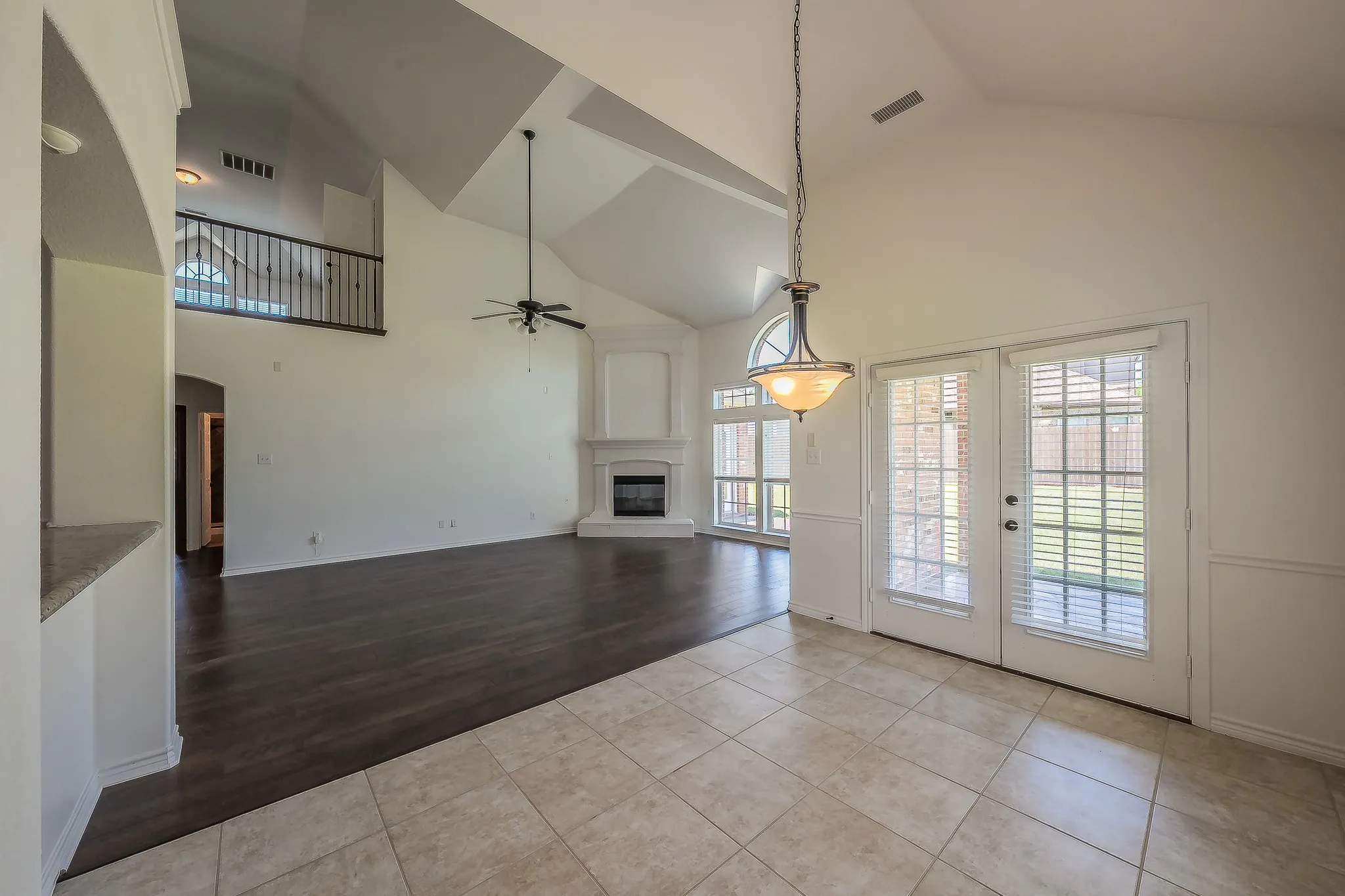 Looking from the breakfast nook into the dramatic two-story family room, showcasing the soaring vaulted ceiling, ceiling fan, fireplace with white surround, arched windows, French doors, and the open staircase landing above.