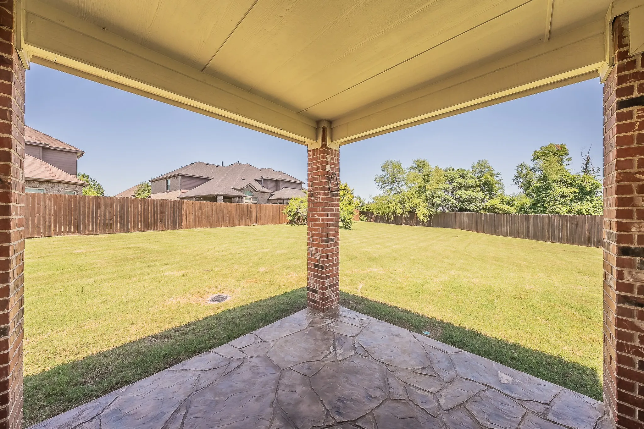 Covered back patio with a stamped concrete floor and brick columns offering a shaded outdoor living area overlooking a large, fully fenced backyard with mature trees — perfect for entertaining.