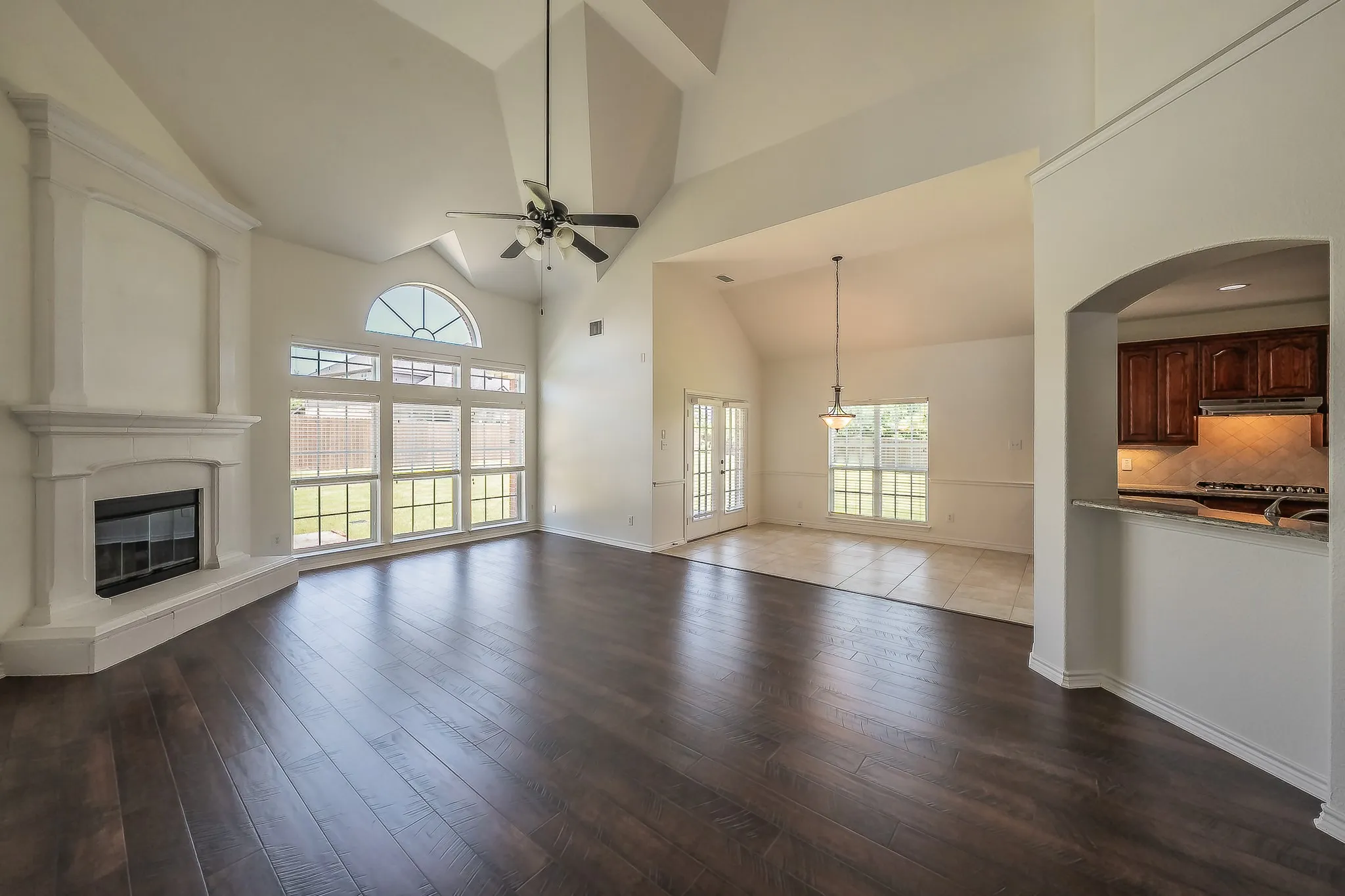 Inviting two-story family room with soaring vaulted ceiling, wall of arched windows, French doors, fireplace with classic white surround and built-in mantel nook, ceiling fan, and gleaming dark hardwood floors.