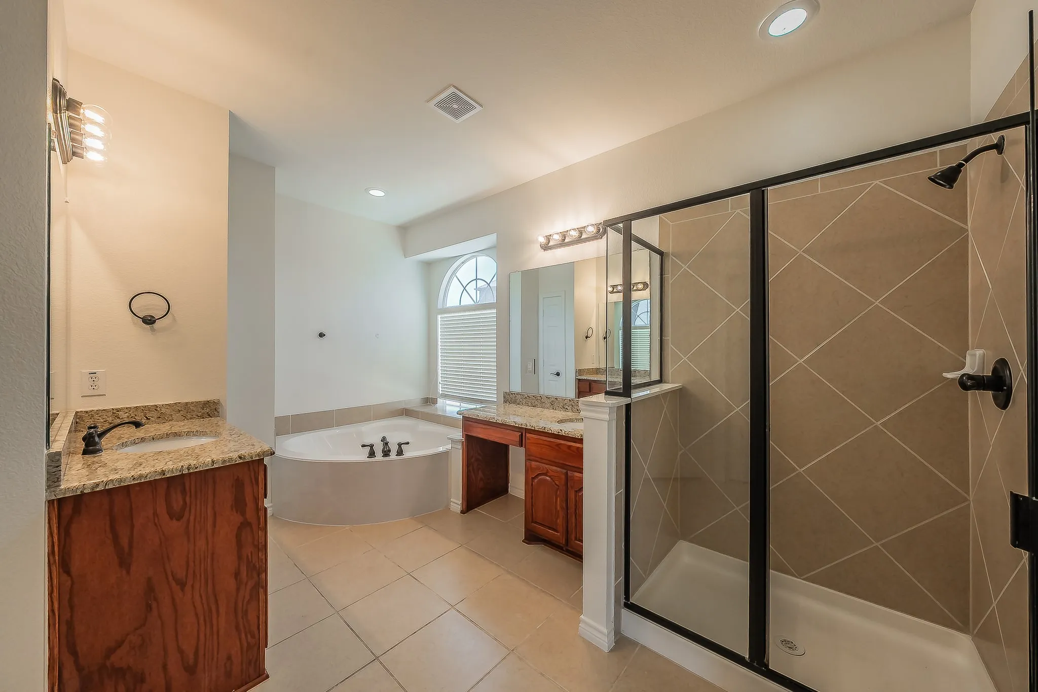Spa-inspired primary ensuite featuring dual granite-topped cherry vanities, large framed mirrors, a corner soaking tub beneath an arched window, large tile walk-in shower with glass enclosure, separate water closet, and tile flooring with oil-rubbed bronze fixtures throughout.