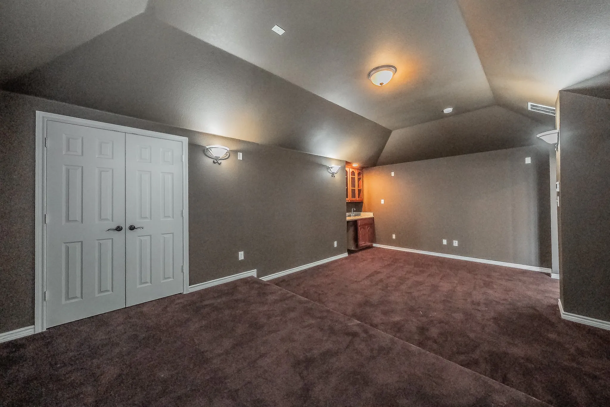 Wide view of the media/bonus room looking toward the built-in wet bar and double-door closet, highlighting the expansive footprint, brand new dark carpet, tiered platform, and ambient sconce lighting — perfect for a home theater, playroom, or game room.
