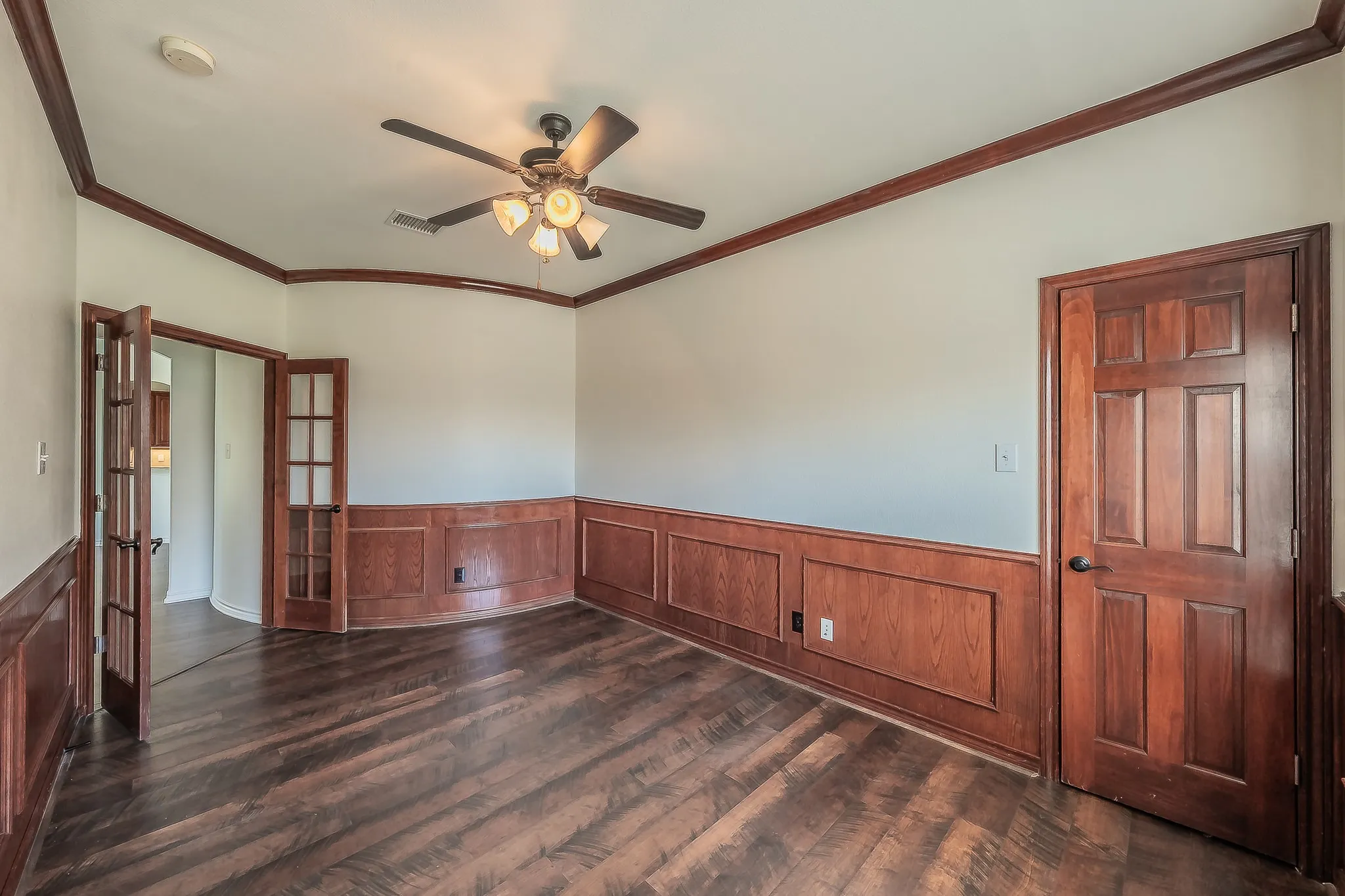 Home office viewed from the French doors showing wood wainscoting wrapping all walls, hardwood flooring, ceiling fan, and a private feel with multiple door entry points.
