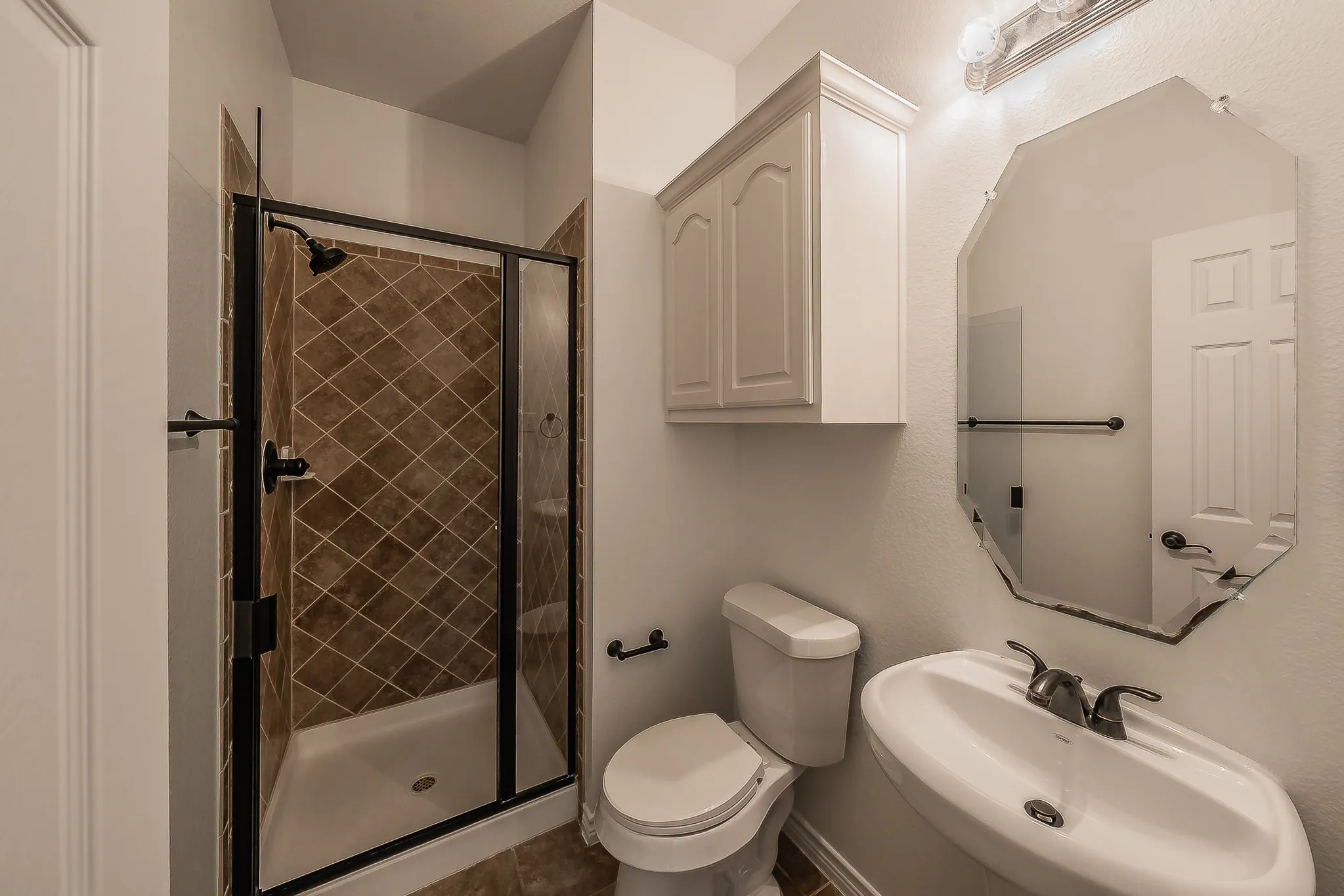Downstairs Bathroom Well-appointed bathroom featuring a tiled walk-in shower with glass door and dark framing, pedestal sink, beveled mirror, white upper cabinet, and oil-rubbed bronze fixtures throughout.