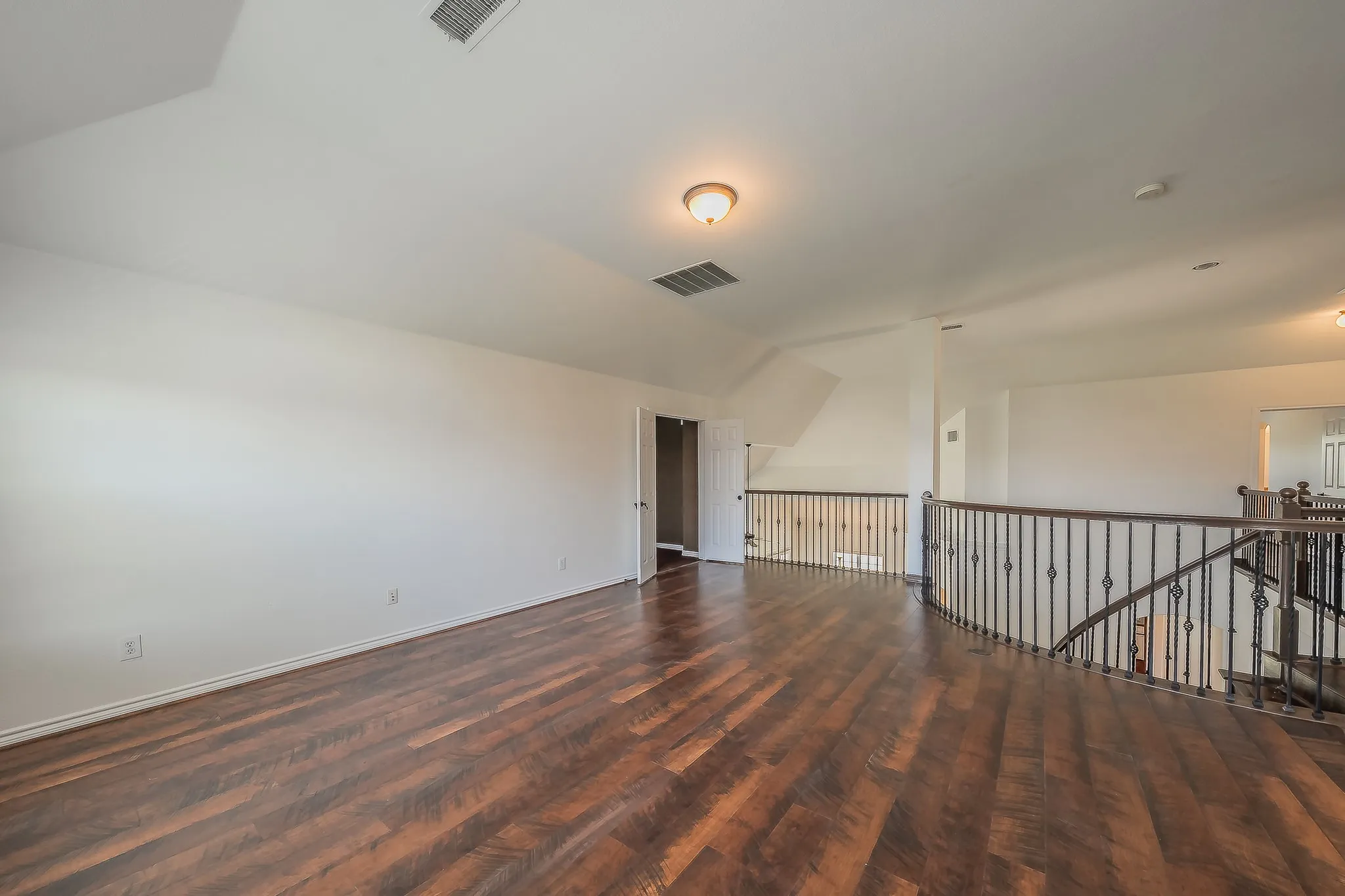 Second-floor hallway viewed from the opposite direction, showing hardwood floors, wrought iron balcony railing, closet access, and open flow connecting the bedroom wing to the loft area.