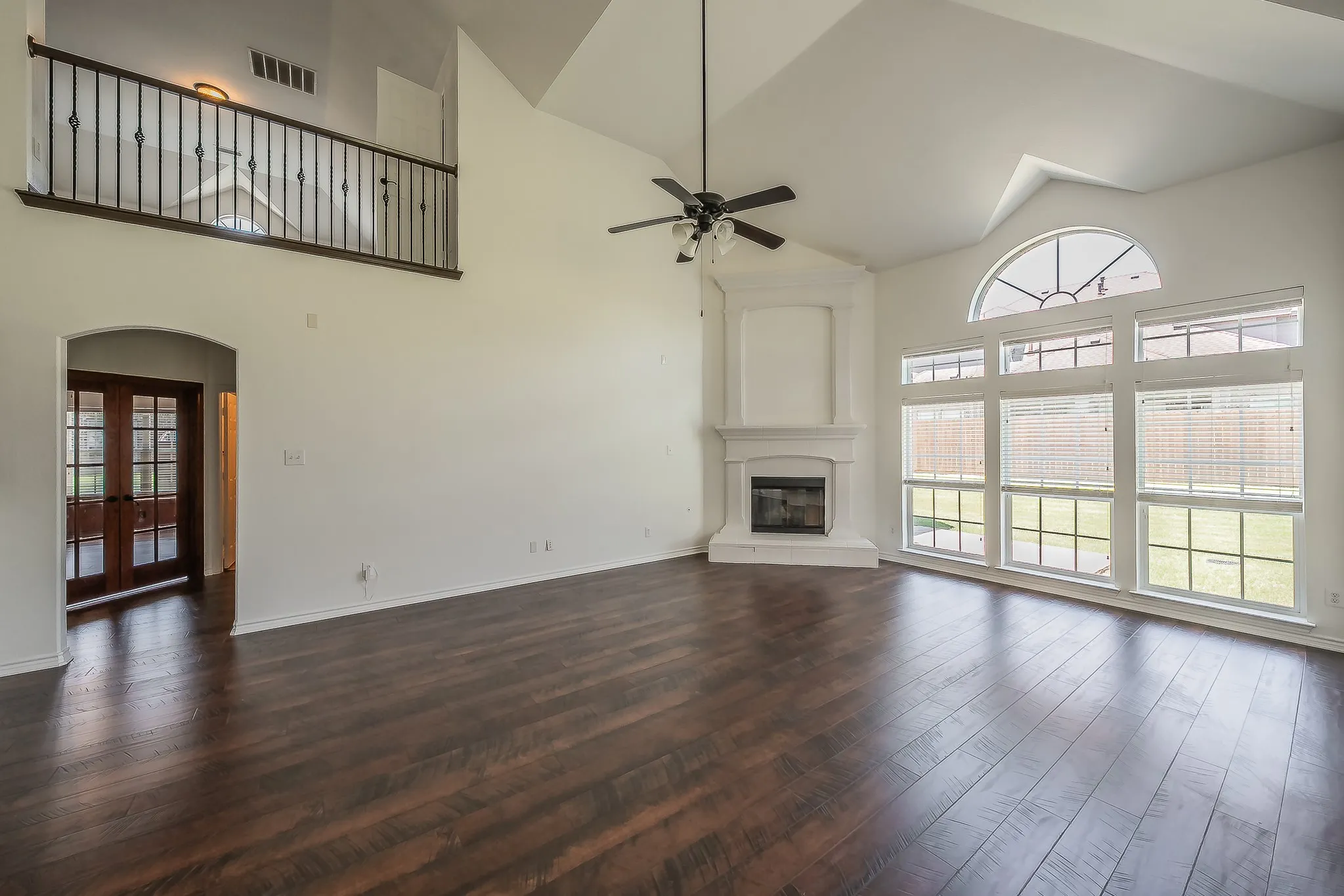 Family room from the far end, highlighting the dramatic wall of arched and picture windows, white corner fireplace with mantel, soaring two-story ceiling, ceiling fan, dark hardwood floors, and view to the French door entry.