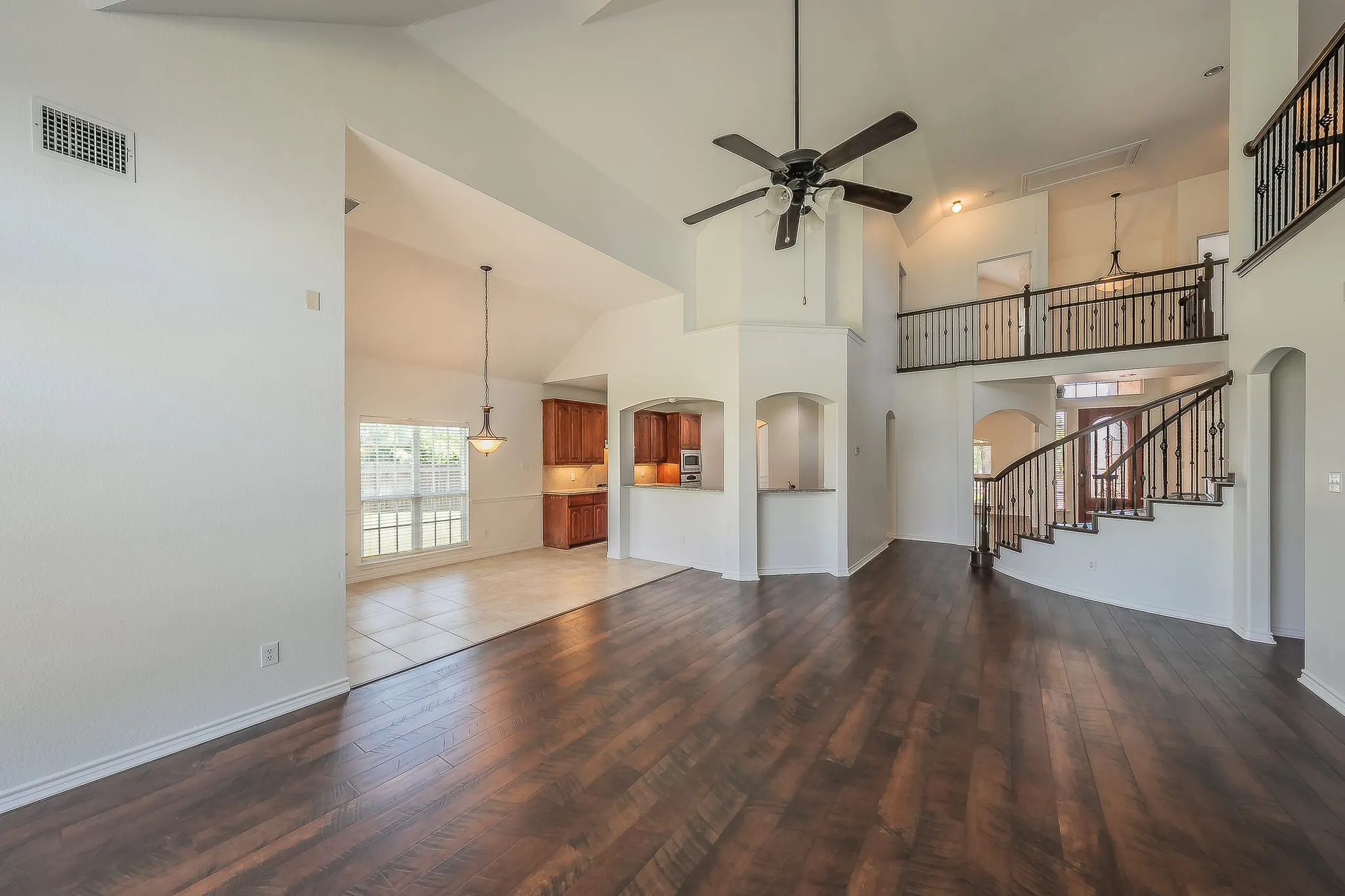 Stunning view of the two-story family room emphasizing the dramatic ceiling height, wrought iron staircase railing on the upper balcony, ceiling fan, and the open connection to the kitchen and nook areas.
