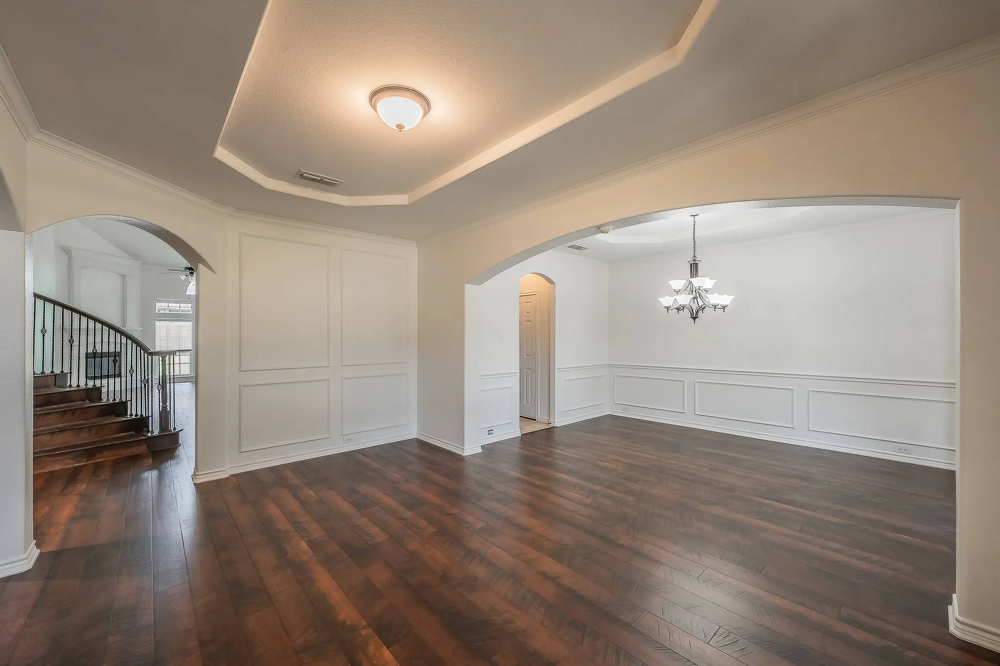 View across the formal living room highlighting the wainscoting accent wall, tray ceiling with flush-mount fixture, arched openings to both the foyer and the formal dining room with chandelier, and polished hardwood floors throughout.