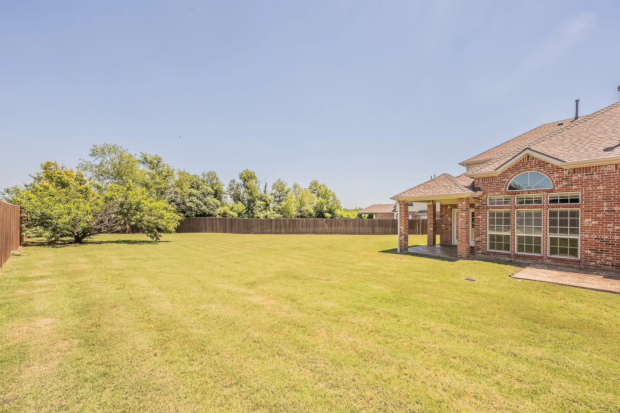 Expansive fully fenced backyard with a lush green lawn, mature landscaping along the fence line, and plenty of room for play, gardening, or a future pool — all under an open Texas sky.