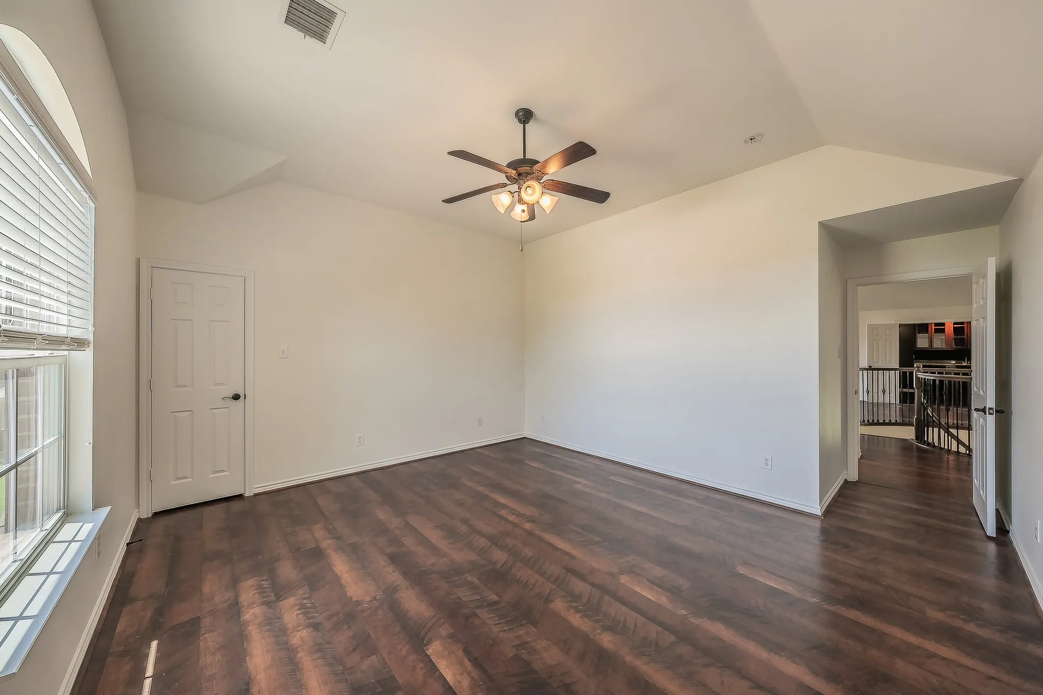 Bright secondary bedroom featuring vaulted ceilings, multiple windows, dark hardwood floors, ceiling fan, and a doorway connecting to the upper hallway with open staircase views beyond.