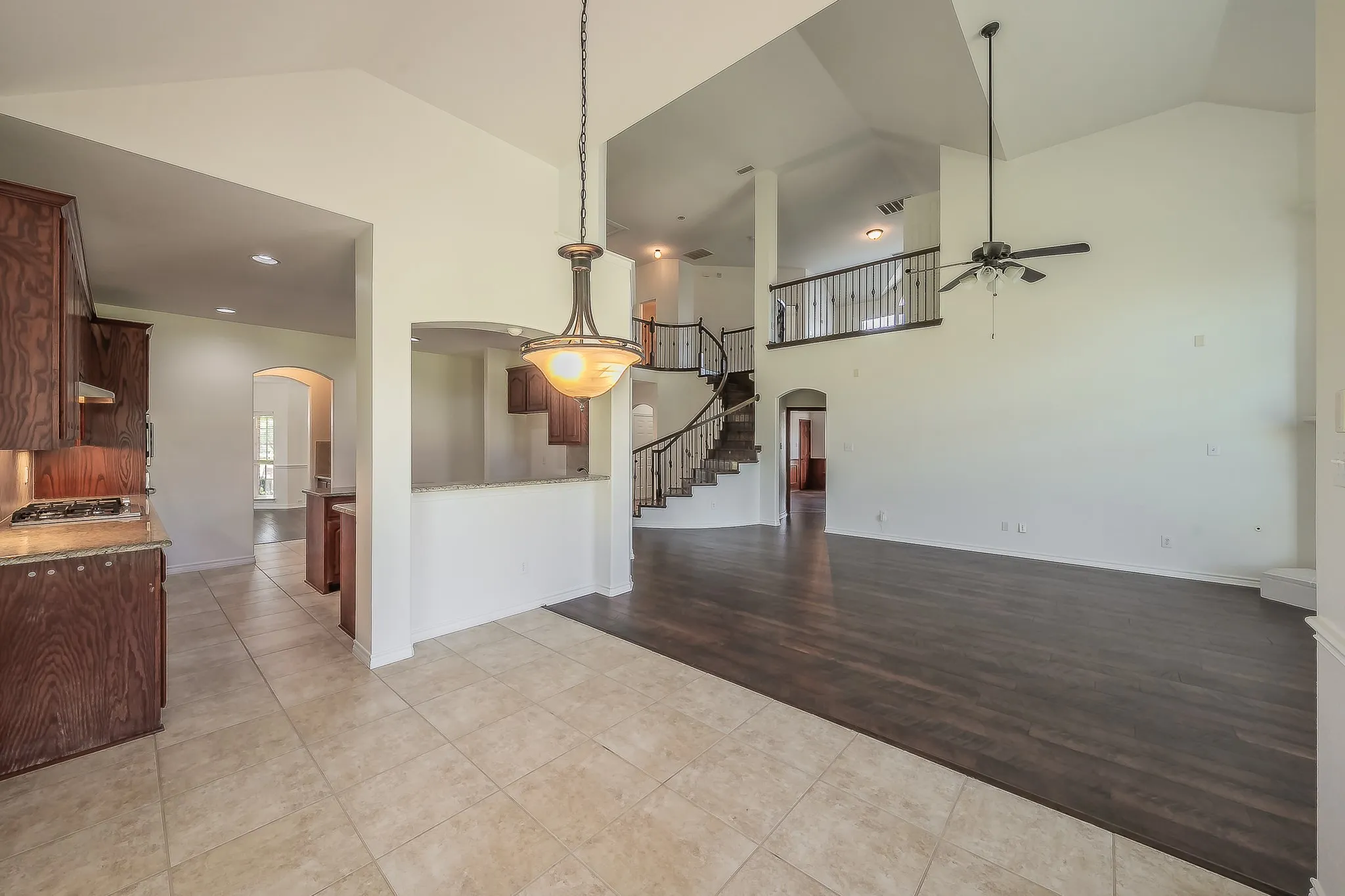 Wide-angle view of the open-plan living area from the kitchen, revealing the two-story family room with soaring ceilings, the curved staircase, wrought iron railing on the upper landing, and the kitchen and breakfast nook seamlessly connected.
