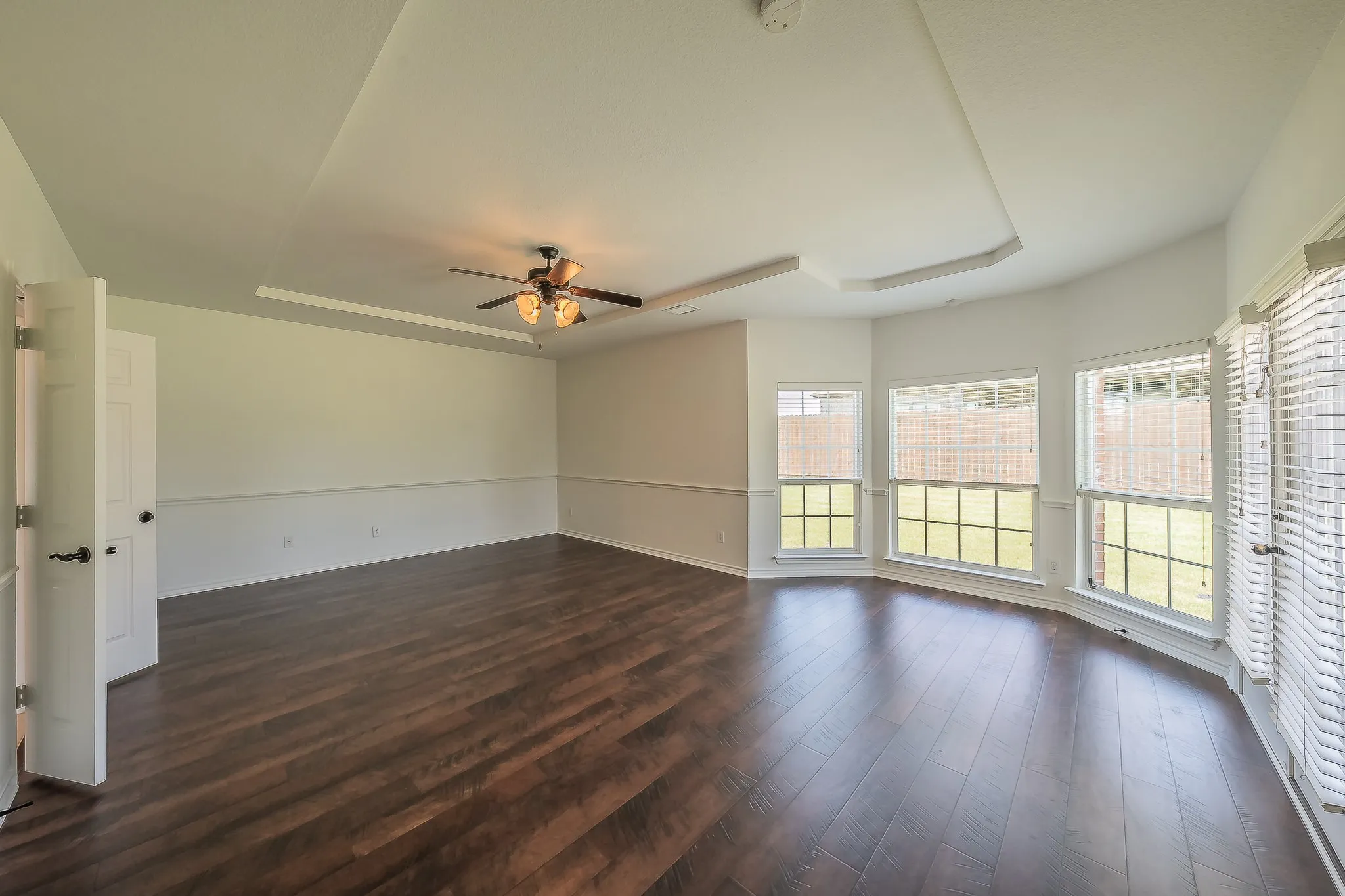 Generously sized primary suite with tray ceiling, ceiling fan, rich hardwood floors, chair-rail wainscoting, and a curved bay window with multiple windows offering abundant natural light.