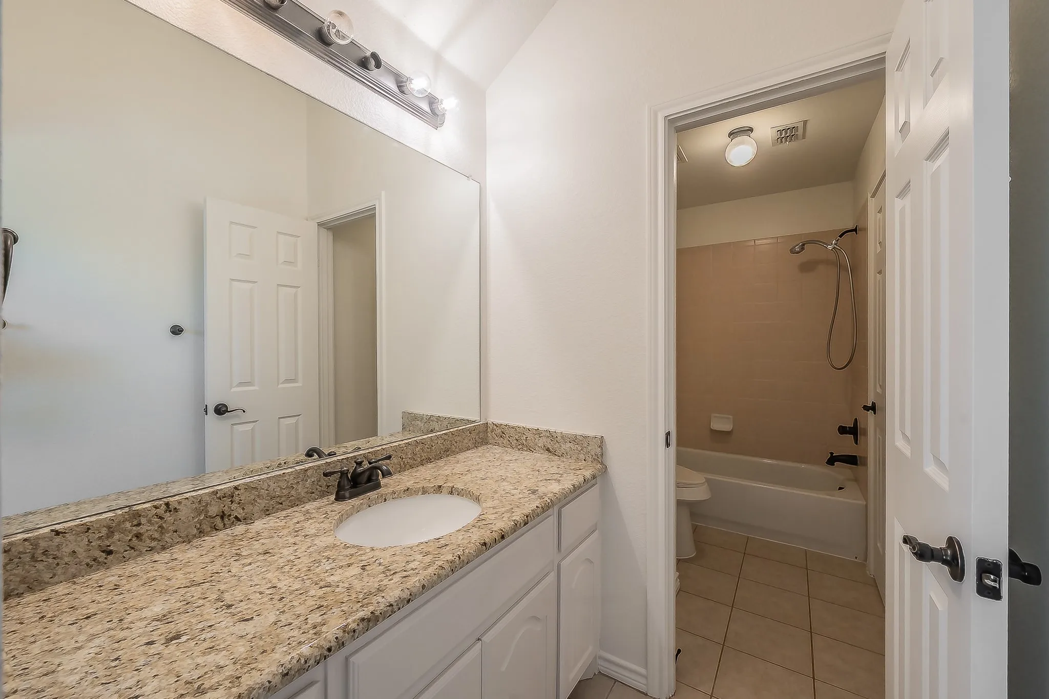 Full secondary ensuite bathroom featuring a granite-topped white vanity, large mirror, oil-rubbed bronze fixtures, tile flooring, and a tub-shower combination with tile surround.
