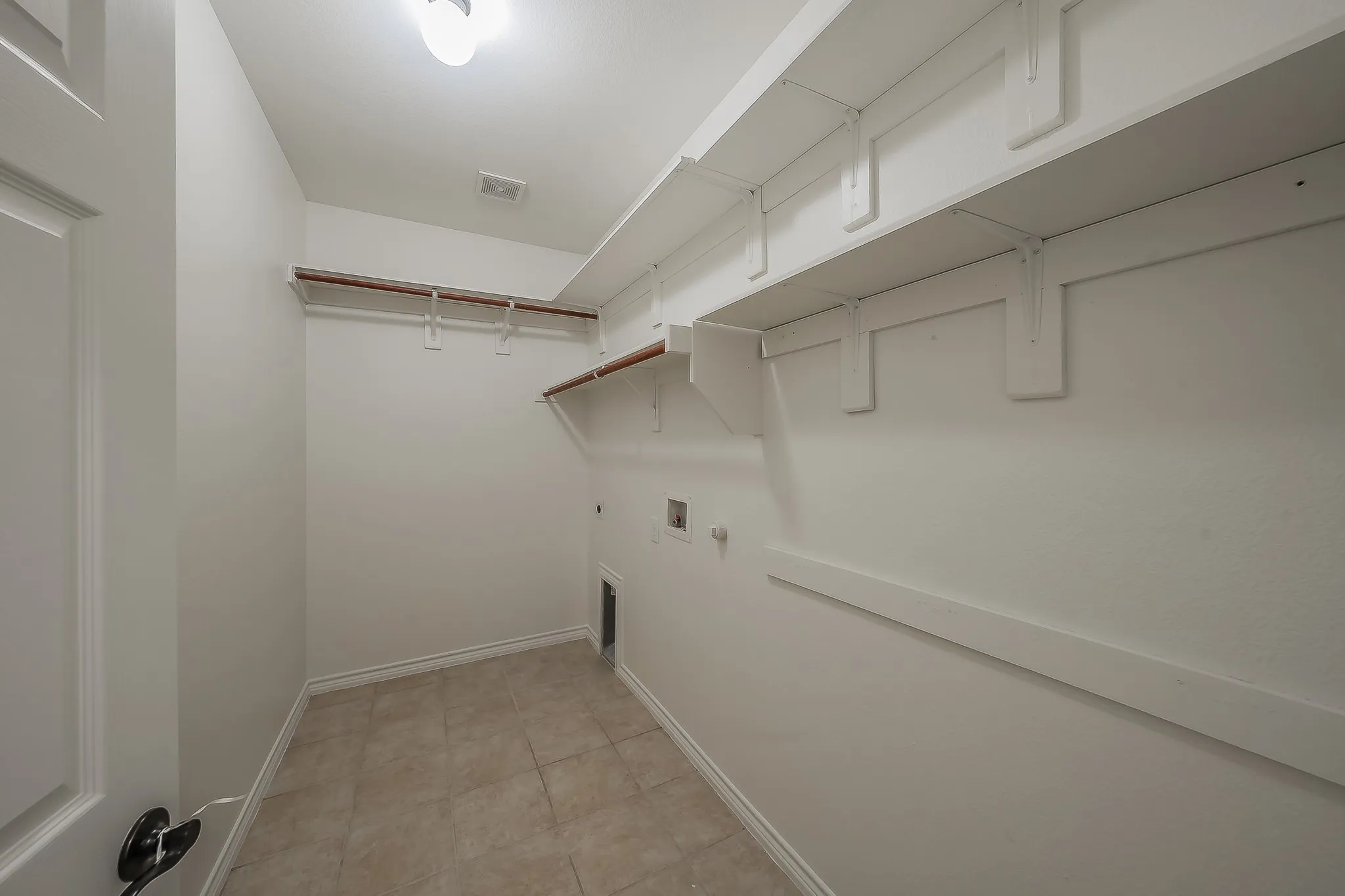 Spacious first-floor laundry room with built-in upper and lower shelving, wood hang rod, tile flooring, washer and dryer connections, and ample storage space.