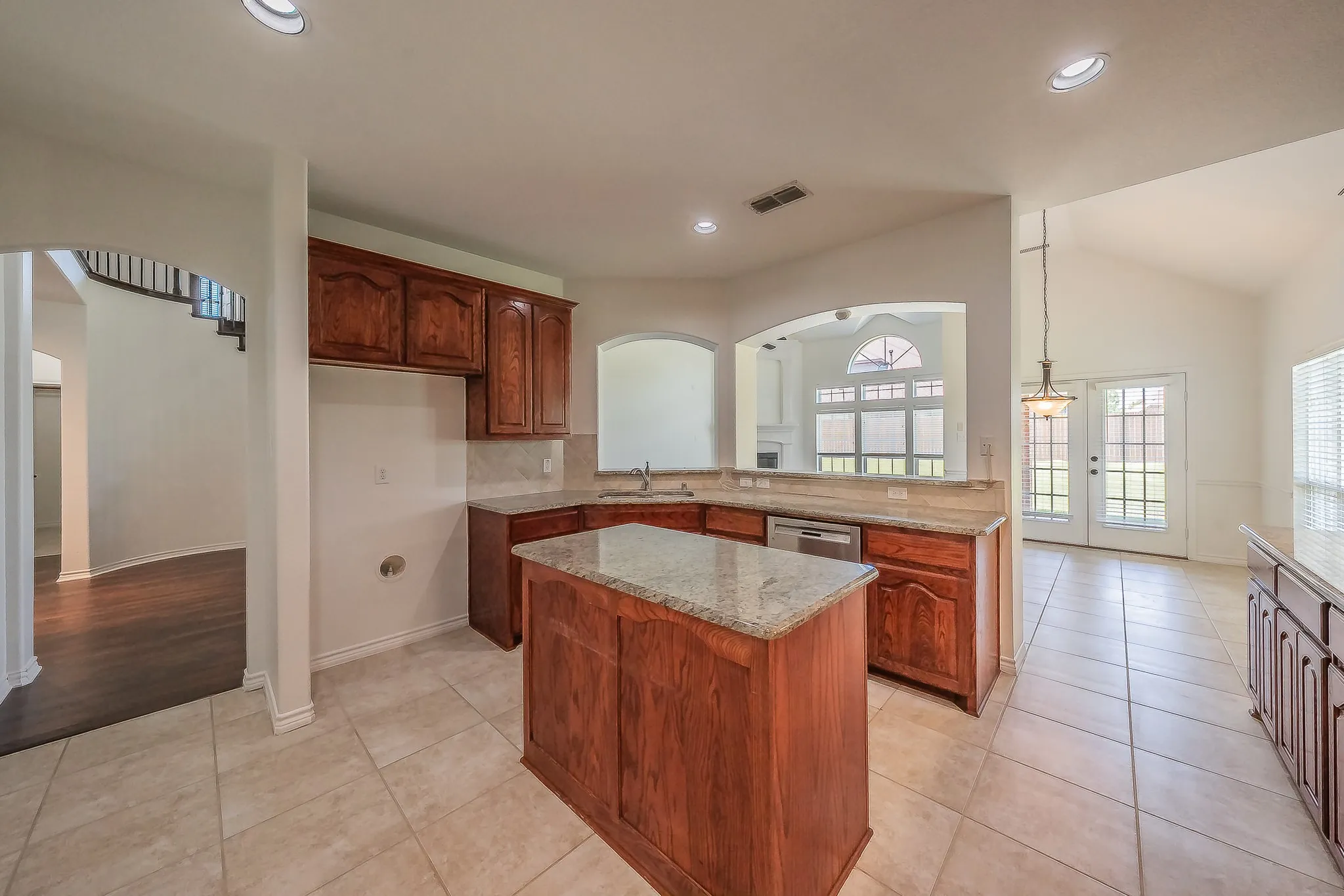 Well-appointed kitchen featuring a granite-topped center island, rich cherry cabinetry with arched raised-panel doors, tile backsplash, stainless dishwasher, and arched windows overlooking the family room and breakfast nook with French doors.