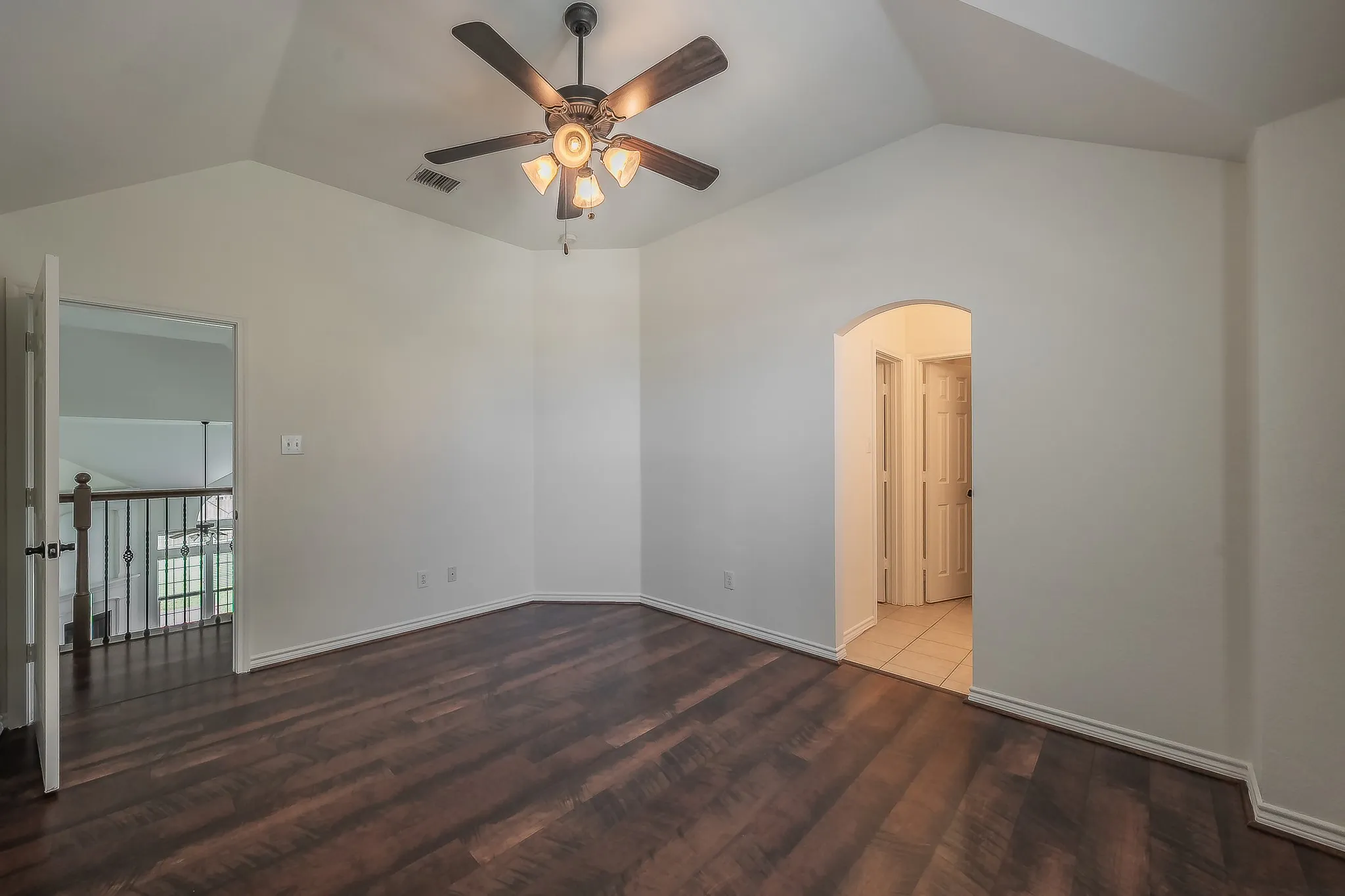 Airy secondary bedroom on the upper level with vaulted ceiling, ceiling fan, dark hardwood floors, and an arched doorway leading to the private ensuite bath with tile flooring.