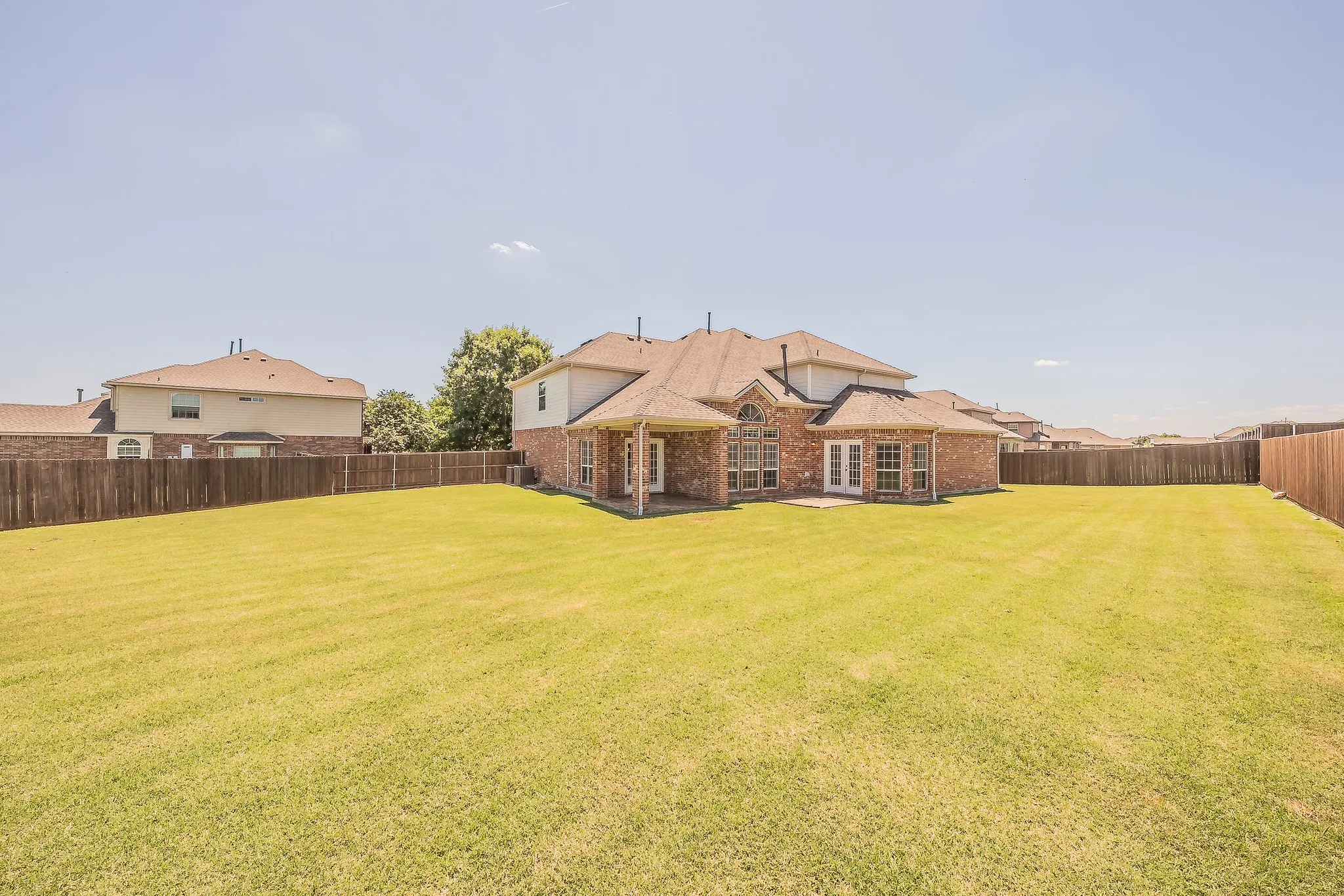 Expansive fully fenced backyard with a lush green lawn, mature landscaping along the fence line, and plenty of room for play, gardening, or a future pool — all under an open Texas sky.