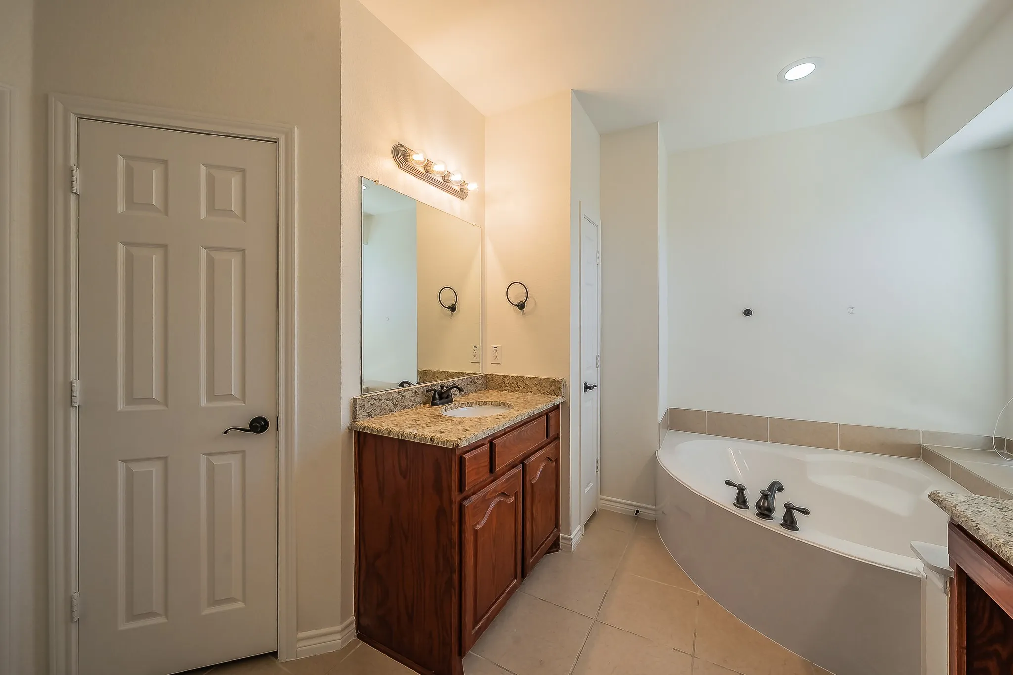 Primary ensuite detail view showing the single vanity with granite countertop, large mirror, oil-rubbed bronze fixtures, and the corner garden soaking tub with tile surround and arched window above.