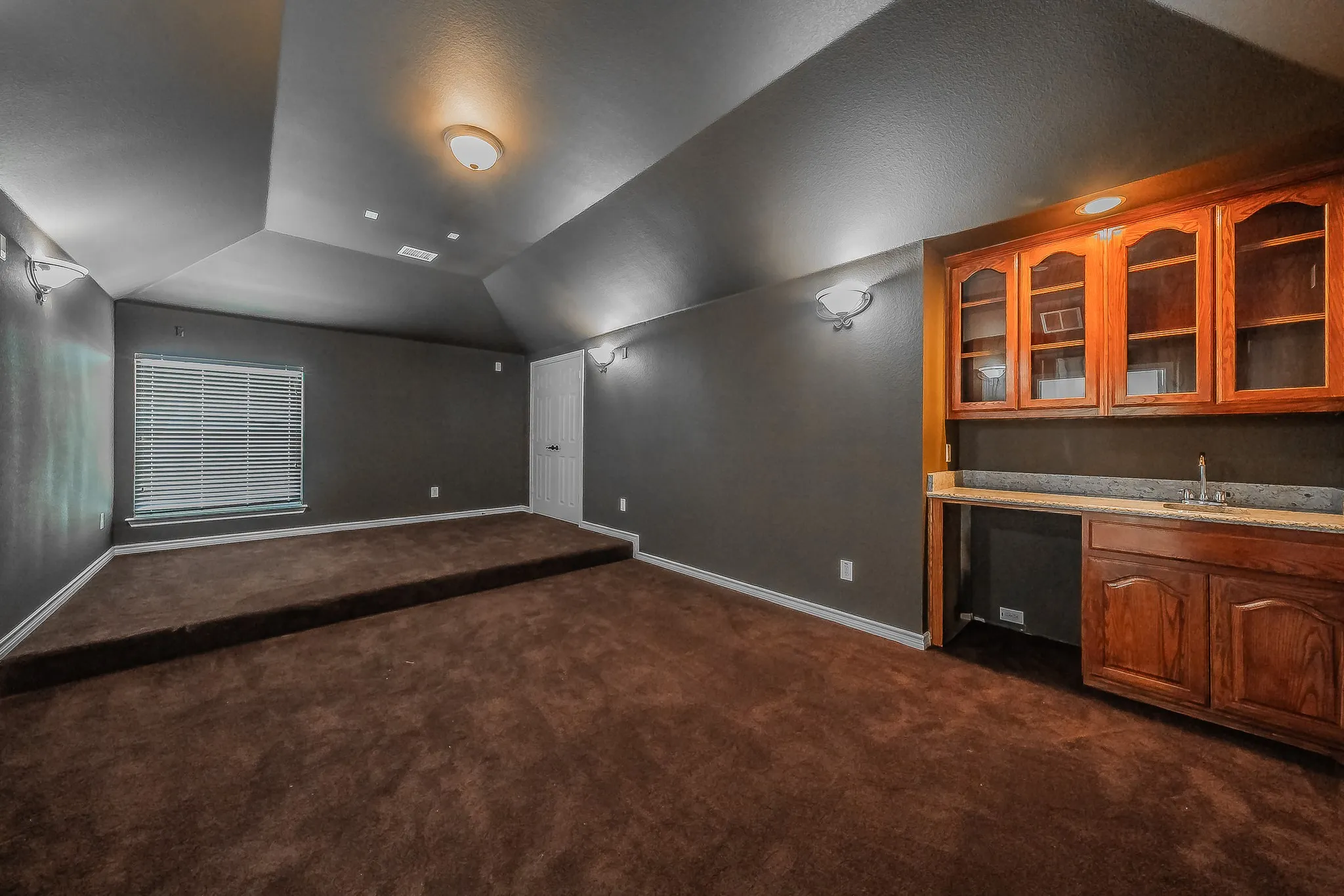 Versatile media room or home theater with dark dramatic walls, tiered carpeted viewing platform, vaulted ceiling, recessed lighting, wall sconces, and a built-in wet bar with cherry glass-front cabinets, granite countertop, and under-counter beverage refrigerator.