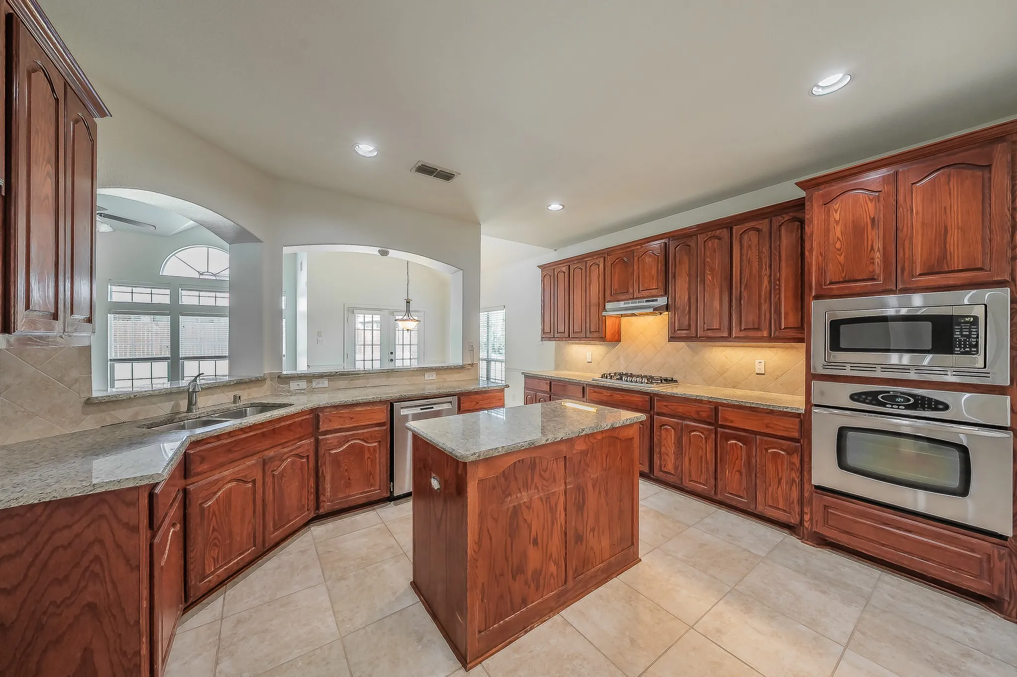 Full kitchen view showcasing extensive cherry cabinetry, granite countertops, center island, tile backsplash, gas cooktop, stainless built oven and microwave, and arched window pass-through connecting to the family room and breakfast nook.