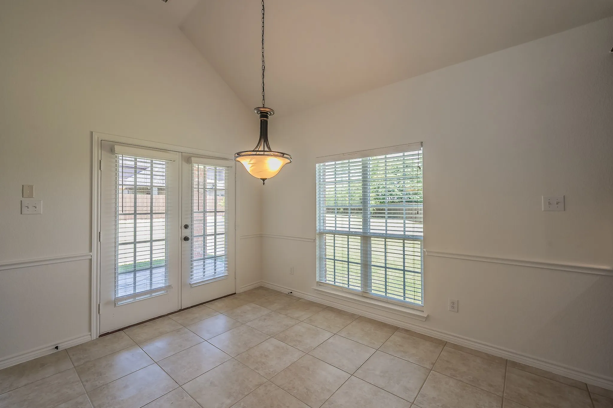 Bright breakfast nook with vaulted ceiling, pendant light, wainscoting, large picture window, and French doors opening to the covered back patio — a cheerful, light-filled space for everyday dining.
