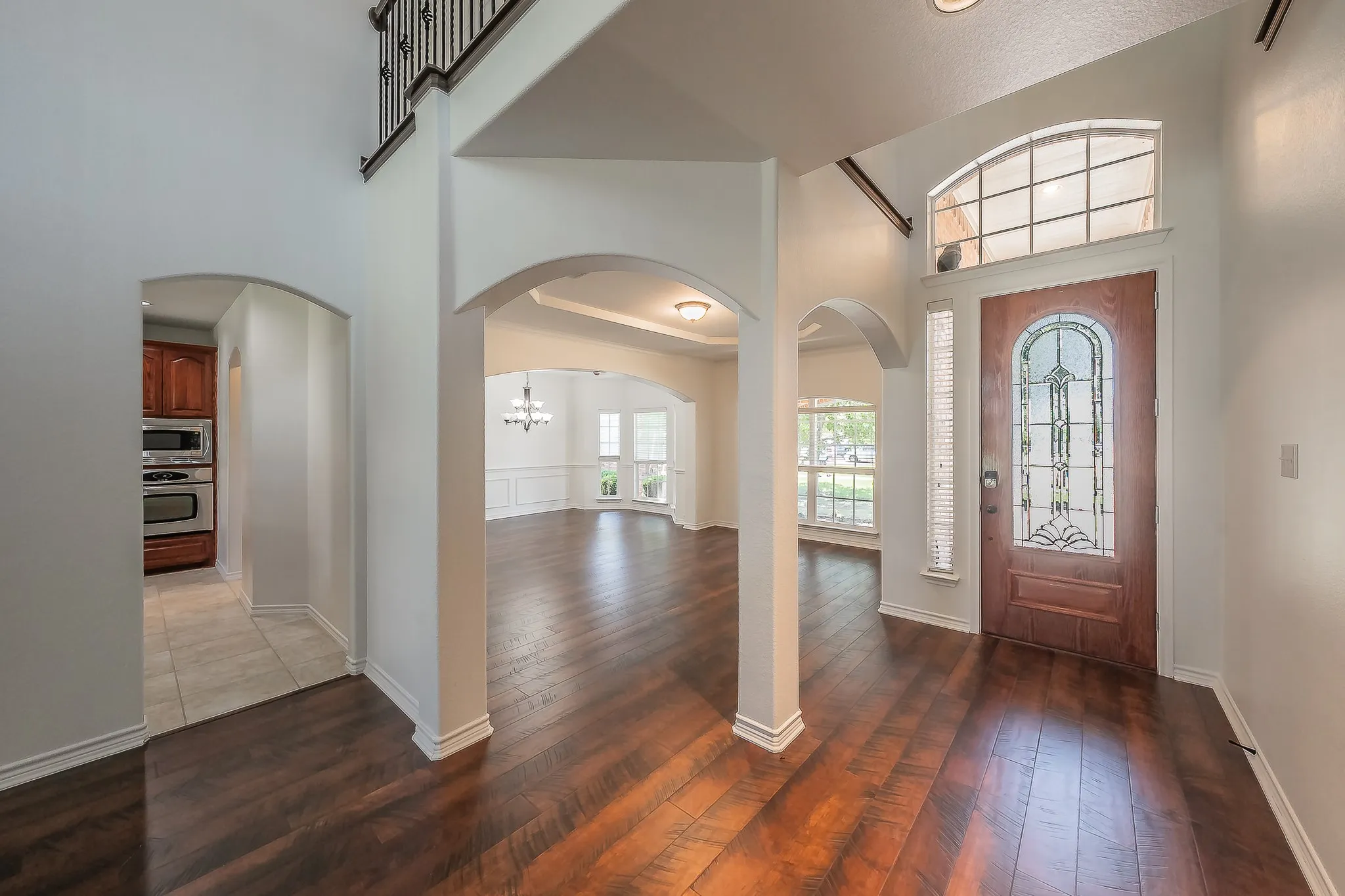 View from the foyer looking toward the front door, showcasing the custom stained-glass front door with sidelite, soaring two-story ceilings, multiple arched doorways connecting to the formal rooms and kitchen, and gleaming hardwood floors throughout.
