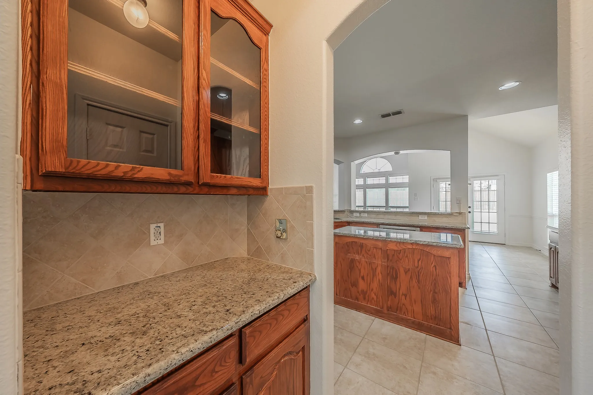 Butler's pantry with cherry glass-front upper cabinets, granite countertop, tile backsplash, and a pass-through view into the kitchen island and breakfast area with French doors and family room beyond.