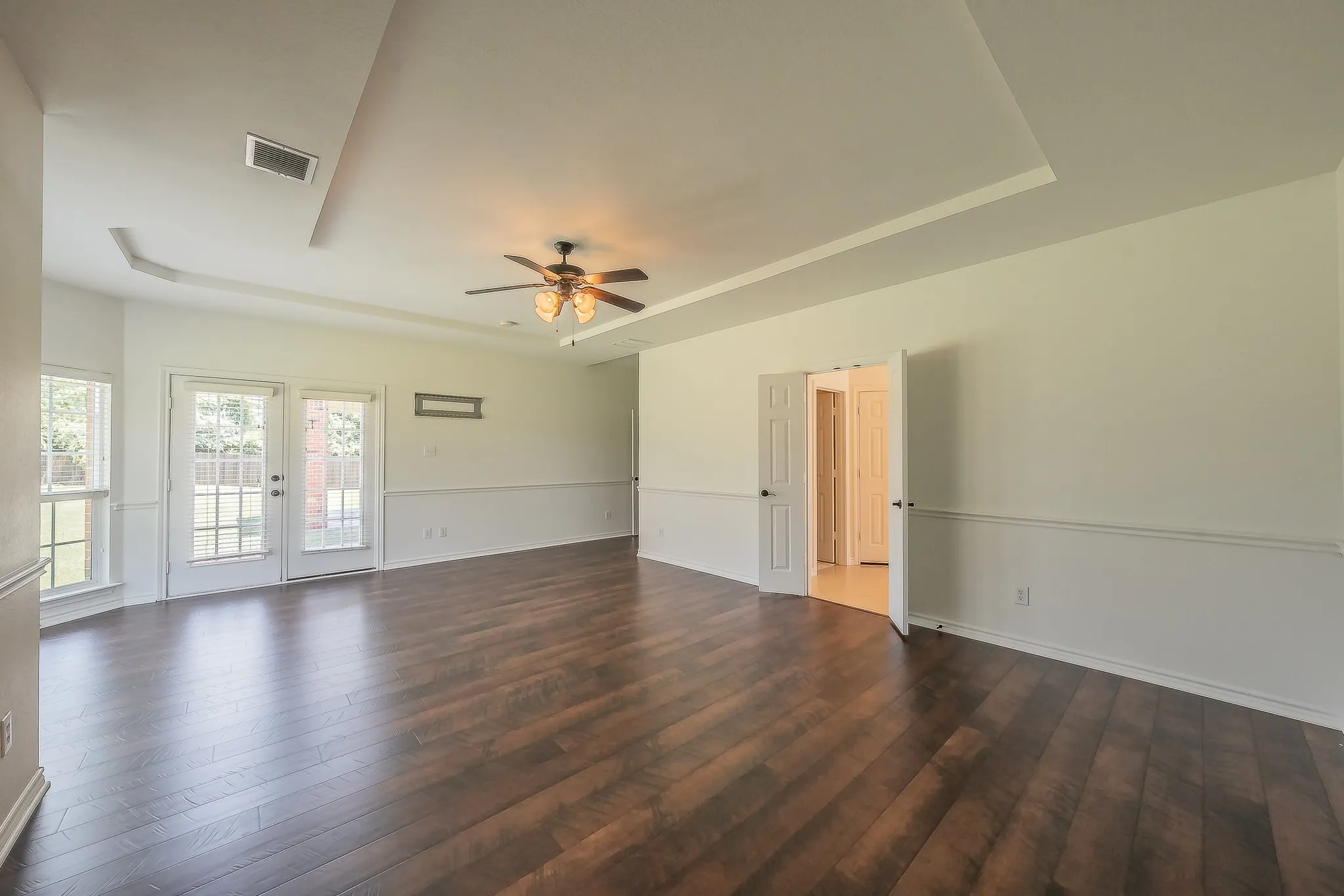 Primary bedroom showcasing French doors opening to a private balcony or exit, ceiling fan, tray ceiling, hardwood floors, and wainscoting — spacious enough to accommodate a large furniture suite