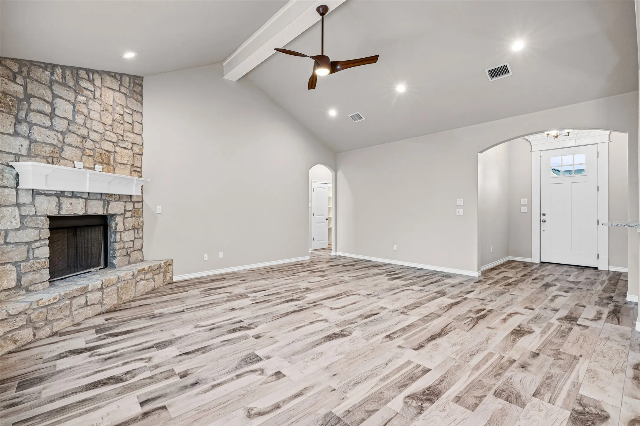 Spacious living area featuring vaulted ceilings with exposed beam, stone-clad fireplace with white mantel, wood-finish flooring, and recessed lighting