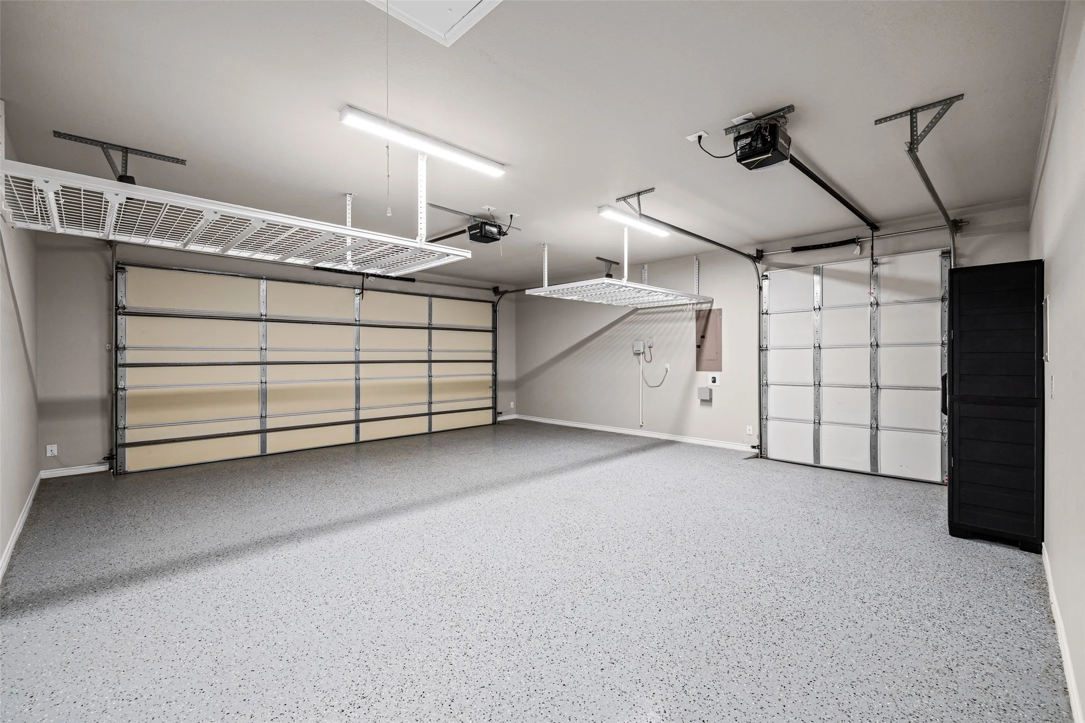 Spacious garage featuring a light gray epoxy floor coating, two overhead storage racks, two automatic garage door openers, and fluorescent lighting