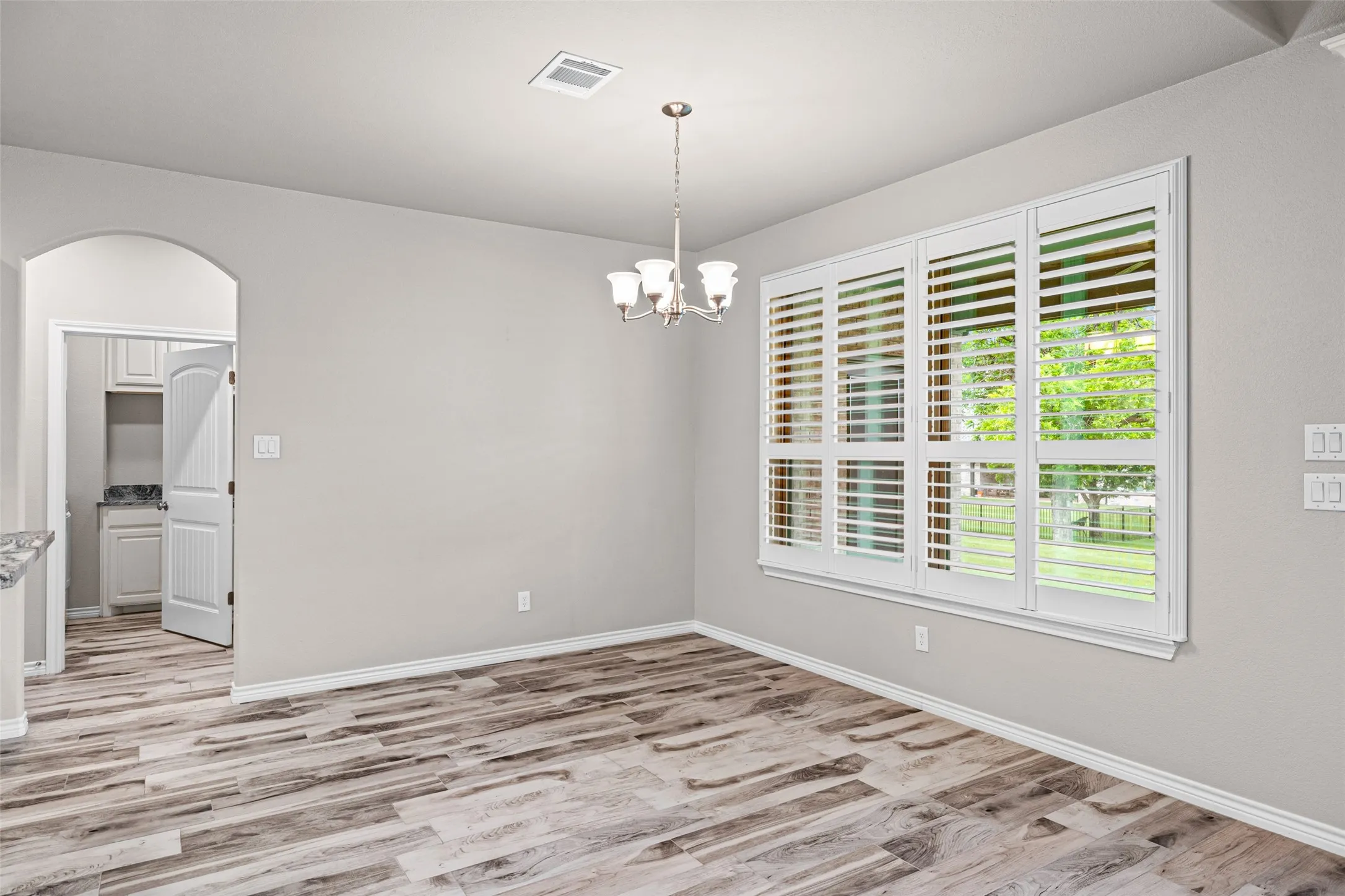 Room featuring wood-finish flooring, a multi-pane window with plantation shutters, a polished nickel chandelier, and an arched entryway