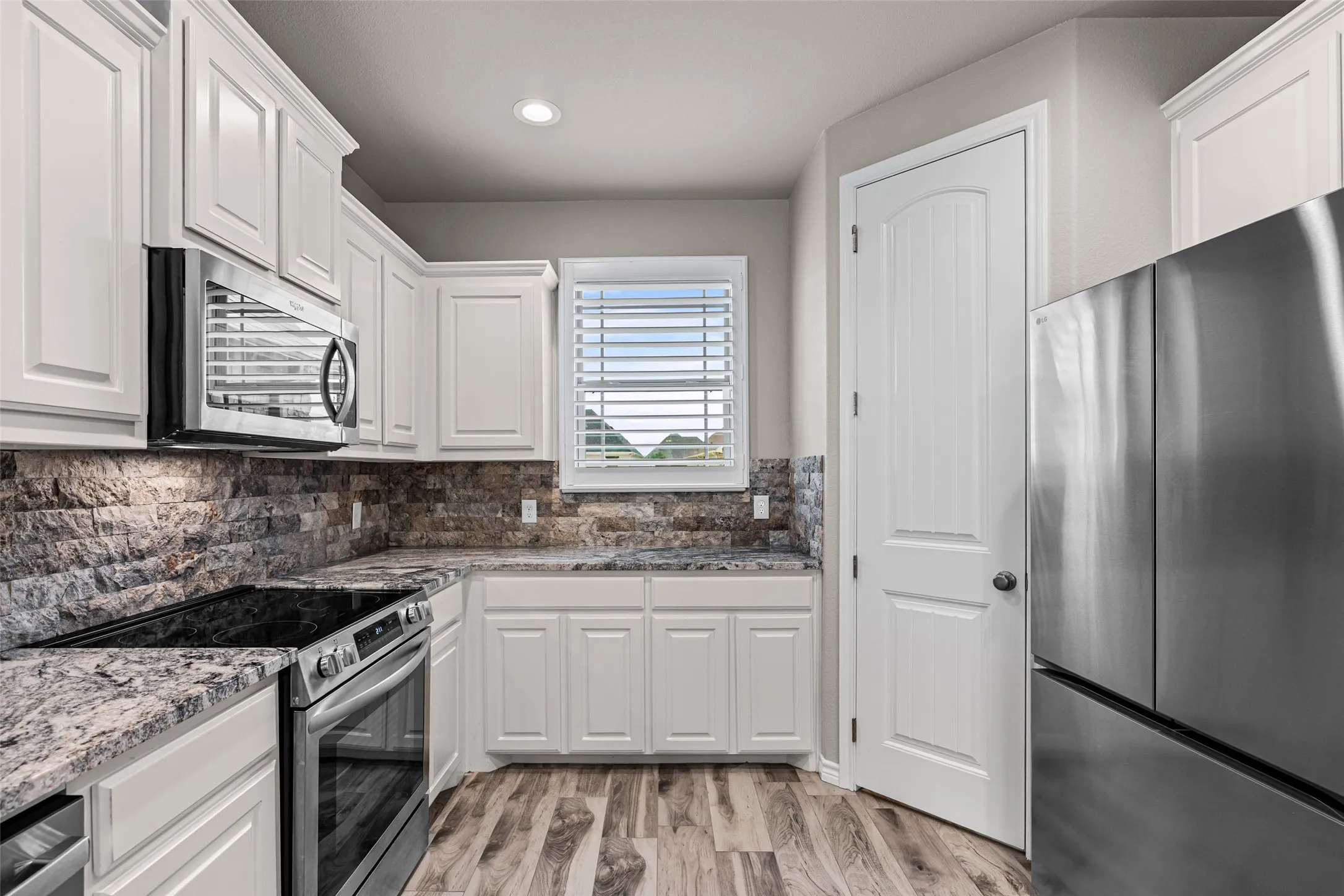 Modern kitchen featuring white cabinetry, stone backsplash, granite-style countertops, stainless steel appliances, and wood-finish flooring