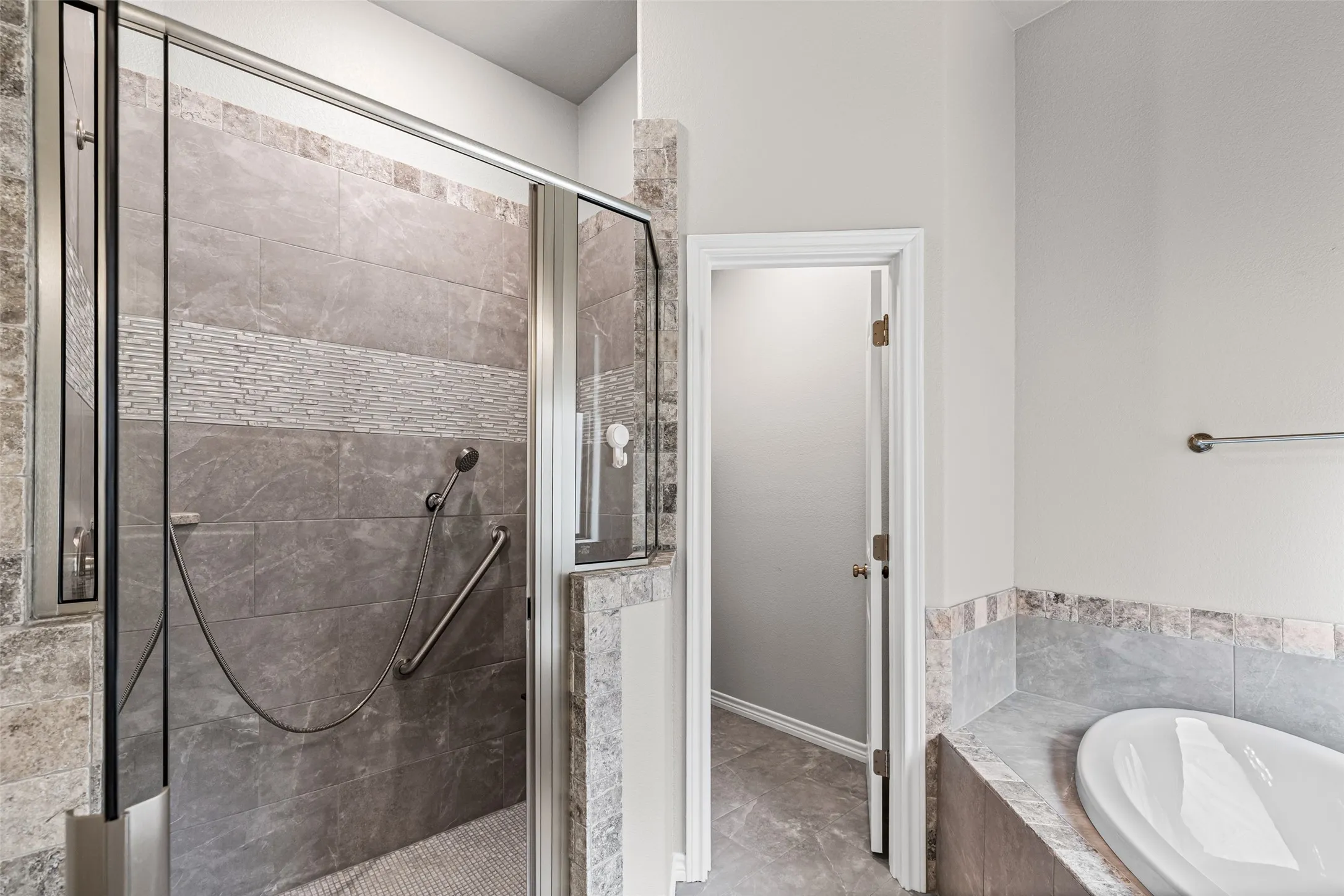 Walk-in shower with glass enclosure, gray tile walls featuring a decorative mosaic band, and a built-in grab bar