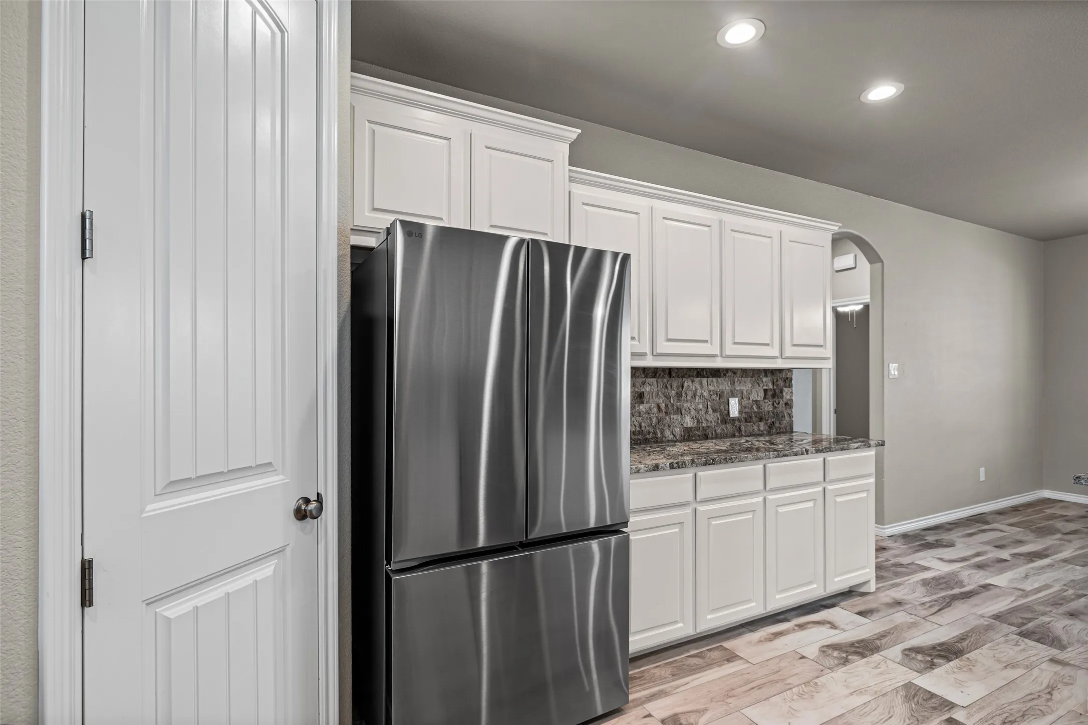 Kitchen area featuring white cabinetry, a stainless steel refrigerator, patterned stone backsplash, and wood-look tile flooring