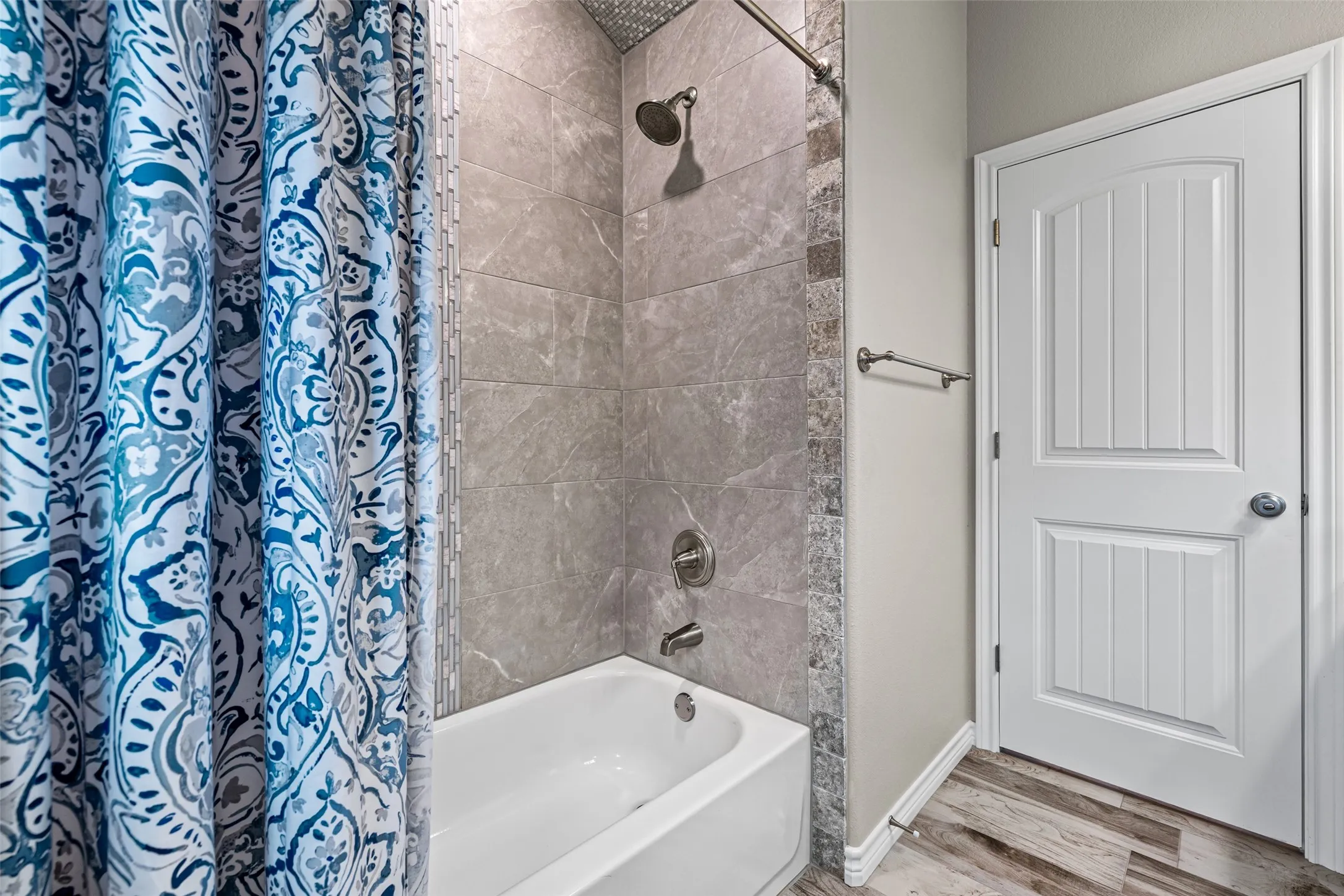 Bathtub and shower combination featuring gray tile surround with decorative mosaic accent, brushed nickel fixtures, and wood-finish flooring