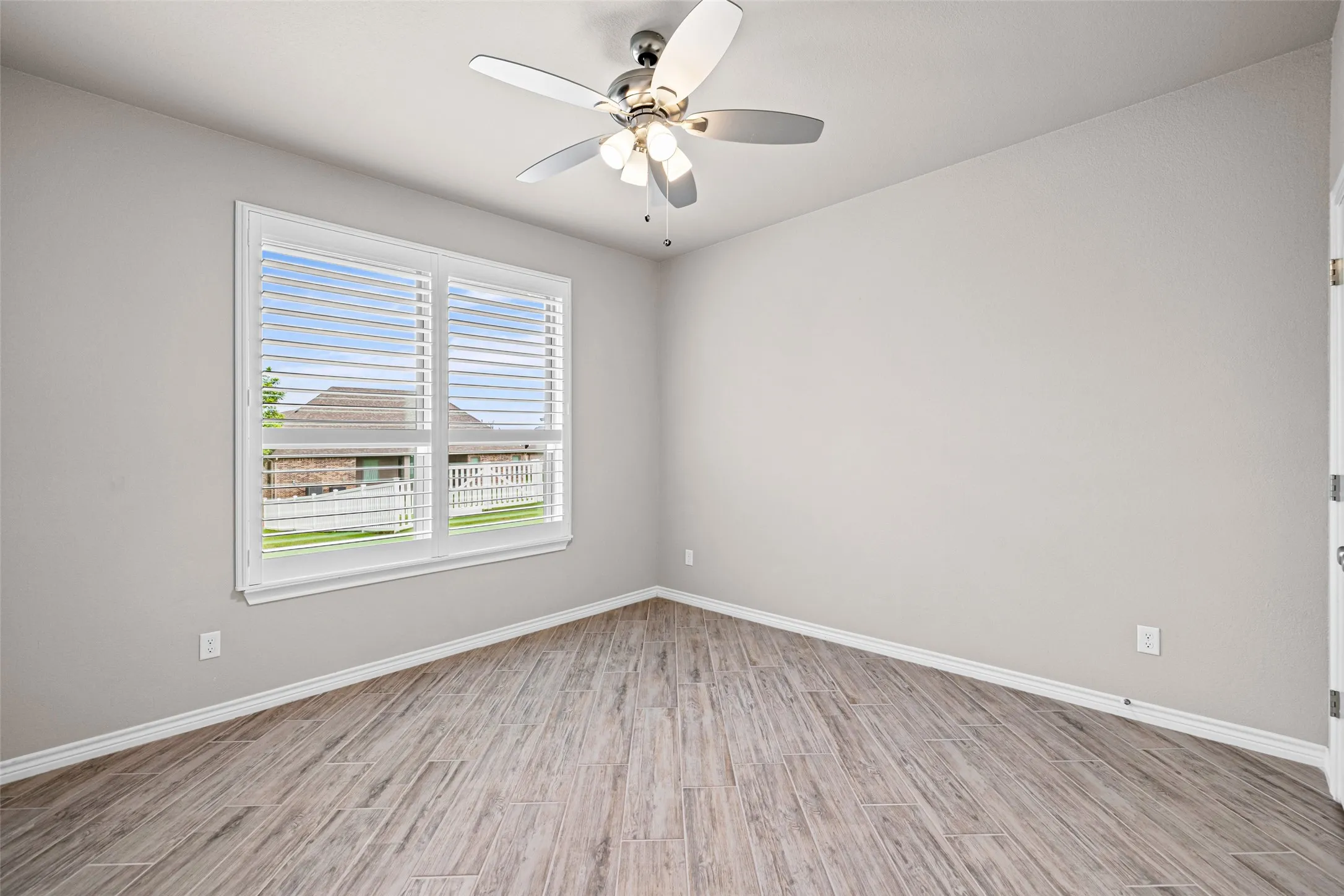 Spacious room featuring wood-finish flooring, a large window with white blinds, and a ceiling fan with integrated lighting