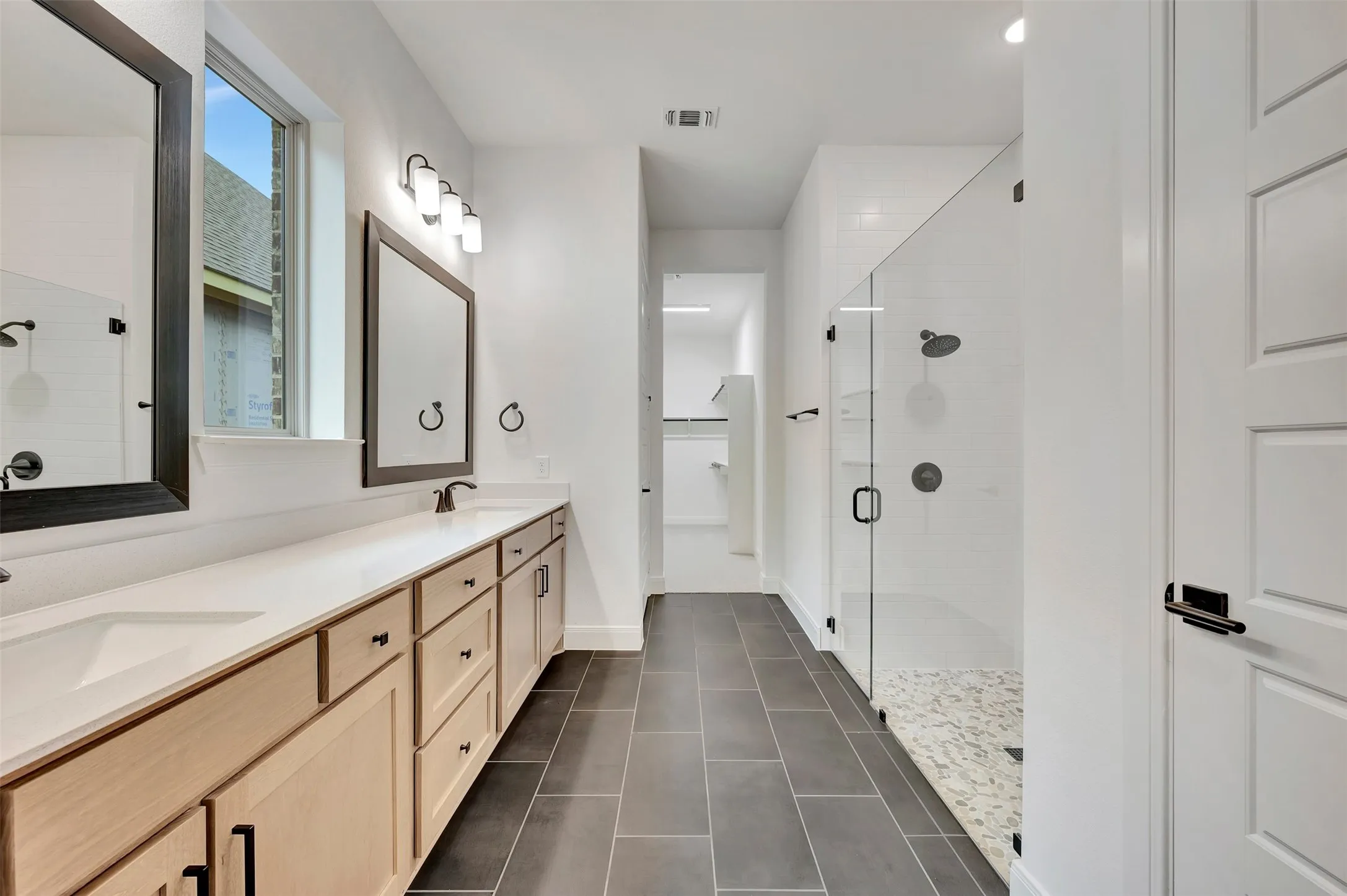 Dual basin vanity with light wood-finish cabinetry, a framed mirror, and a walk-in shower with a glass enclosure and pebble floor