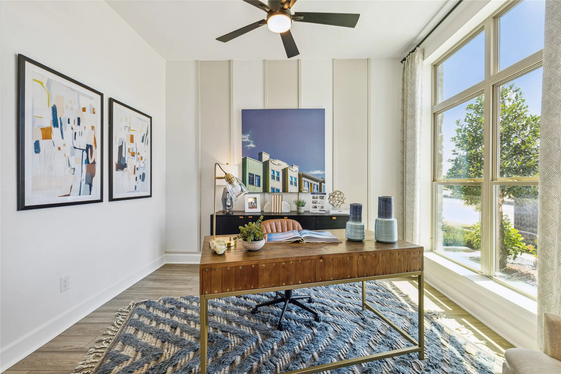 Sophisticated home office featuring a wood-finish desk with brass-tone metal legs, large-scale framed artwork on white walls, and abundant natural light from expansive multi-pane windows