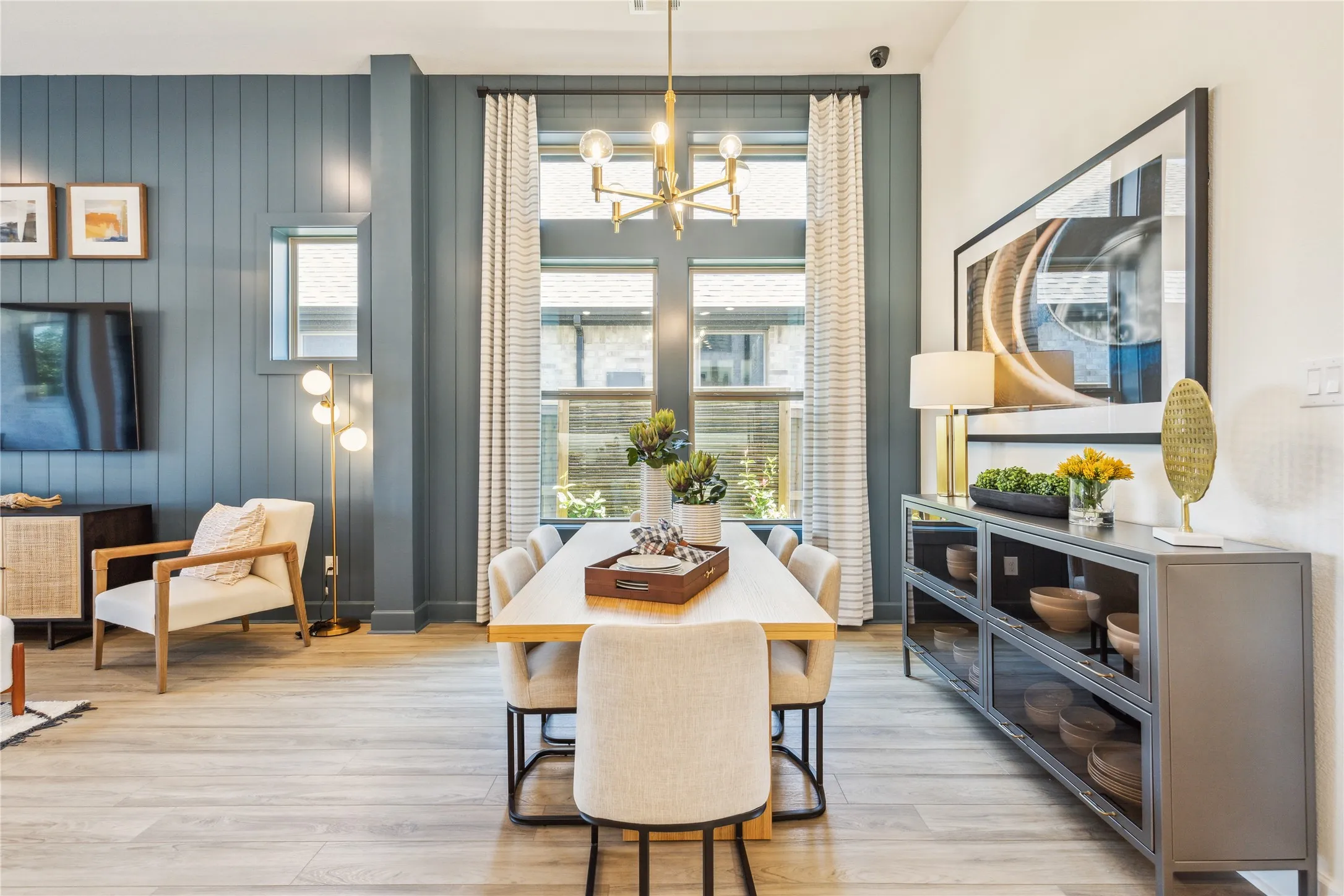 Open-concept living space featuring a wood-finish plank floor, large multi-pane windows with natural light, and a central dining table with upholstered seating