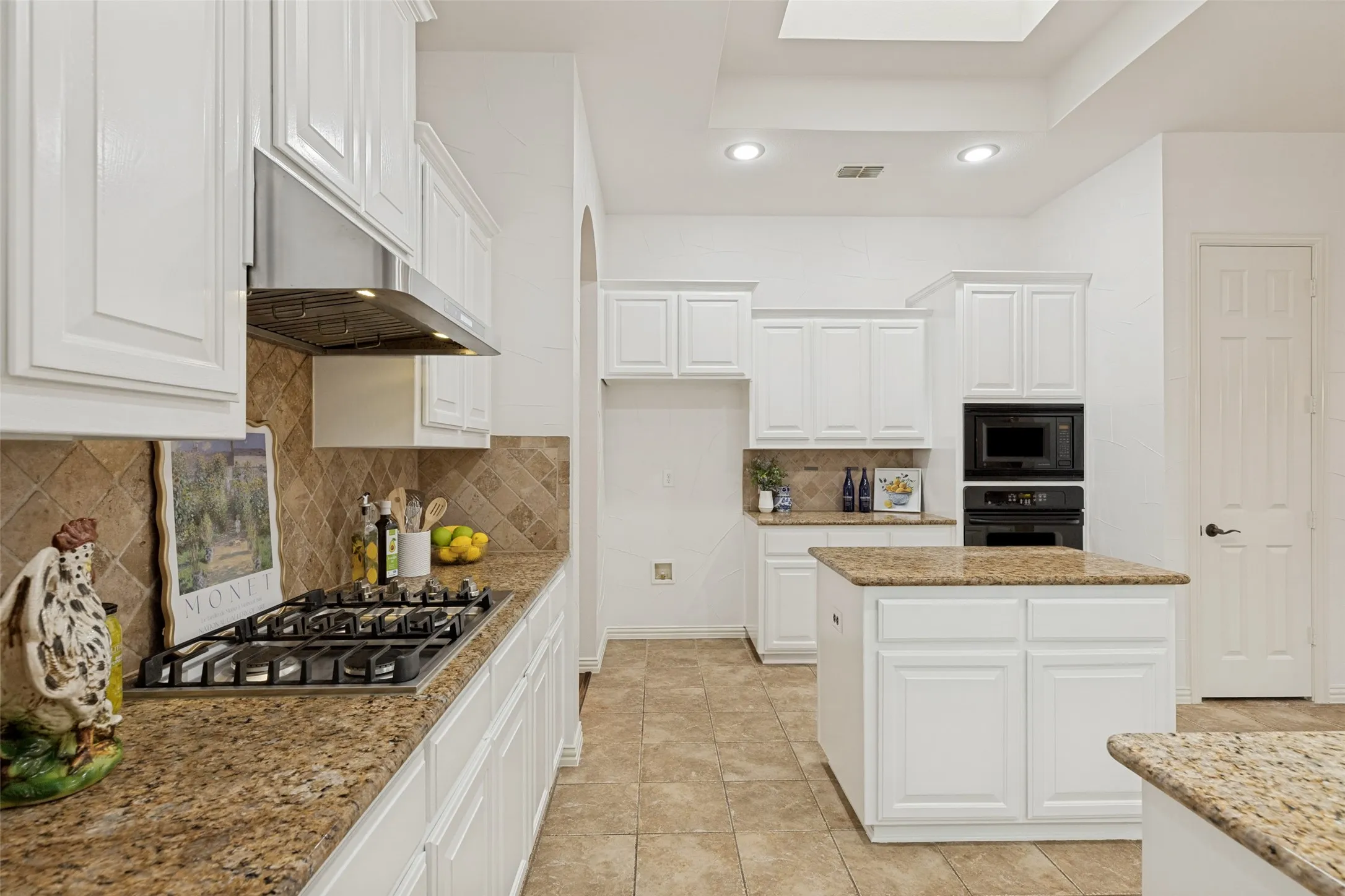 Spacious kitchen featuring white cabinetry, granite countertops, stainless gas cooktop with vent, and a central island with a skylight above.
