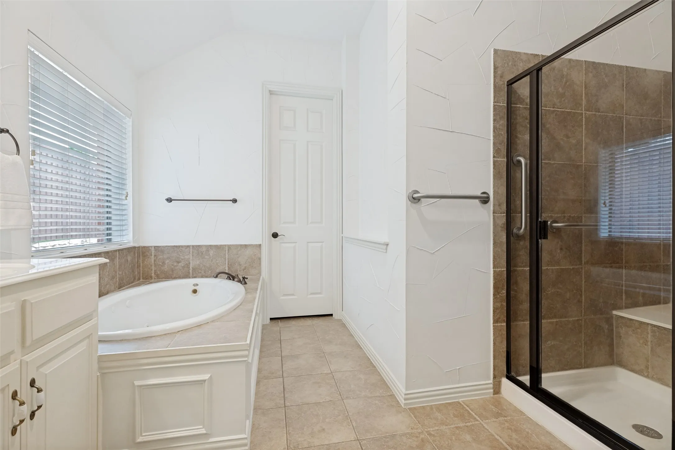 Primary Bathroom featuring a built-in jetted tub with tiled surround, a separate shower with a glass enclosure and bench seating, neutral-toned tile flooring, and a window with blinds.
