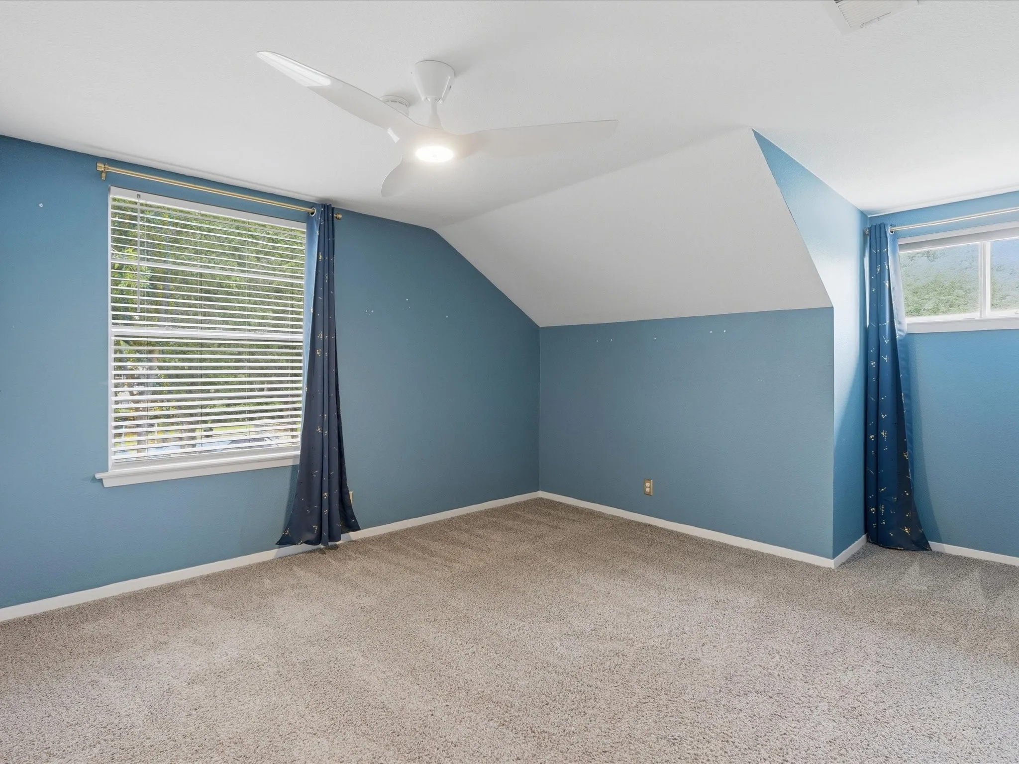 Spacious room featuring sloped ceilings, two windows with blinds, and neutral-toned carpet flooring